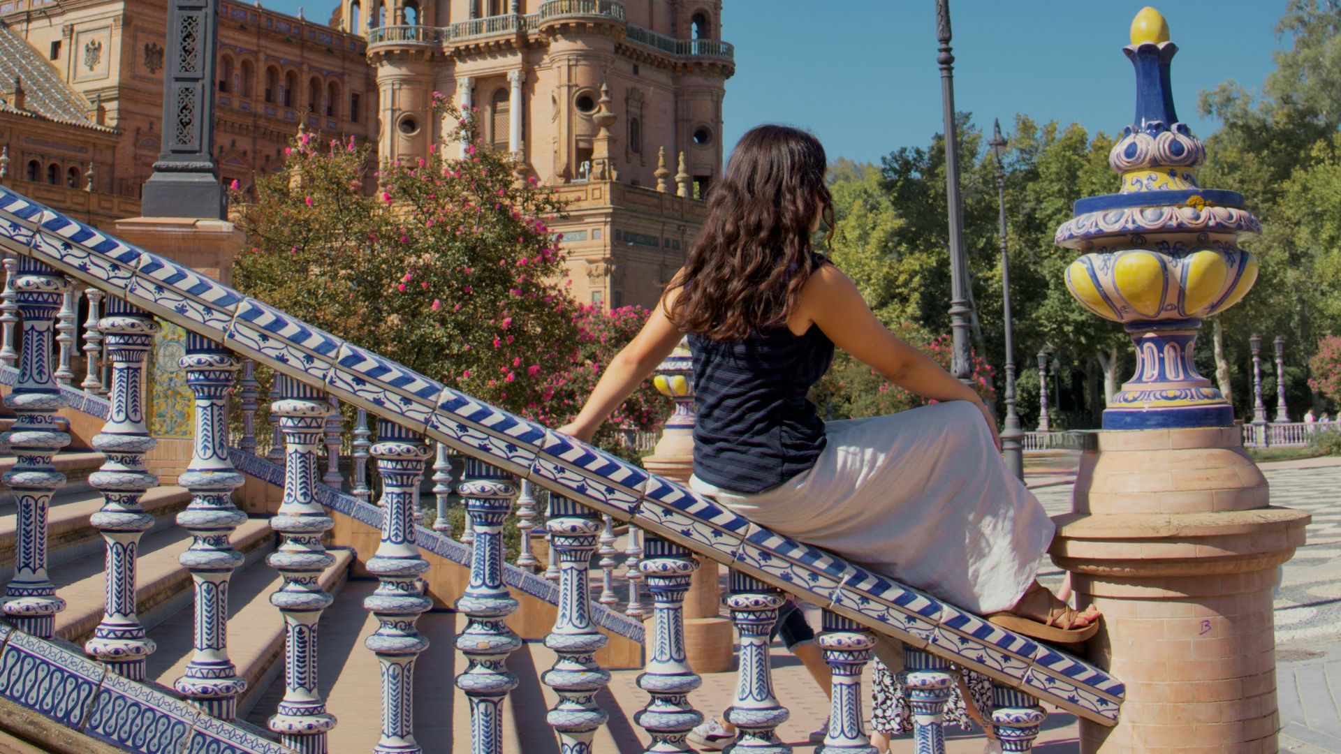 A person sits on an ornate blue and white tiled staircase, looking at a large, historic tower against a clear blue sky.