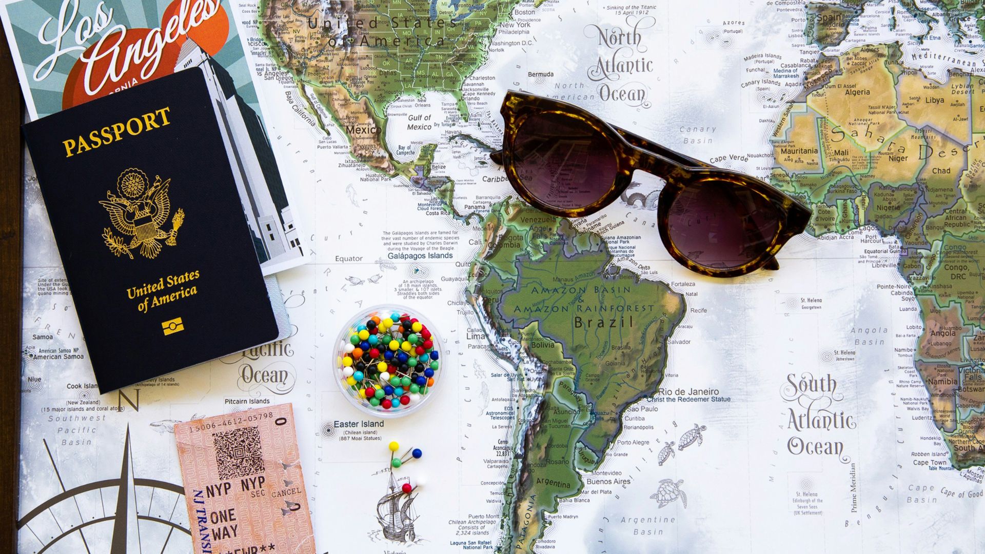 A world map with a passport, sunglasses, a "Los Angeles" postcard, colorful pushpins, and a plane ticket stub laid out for a travel theme.