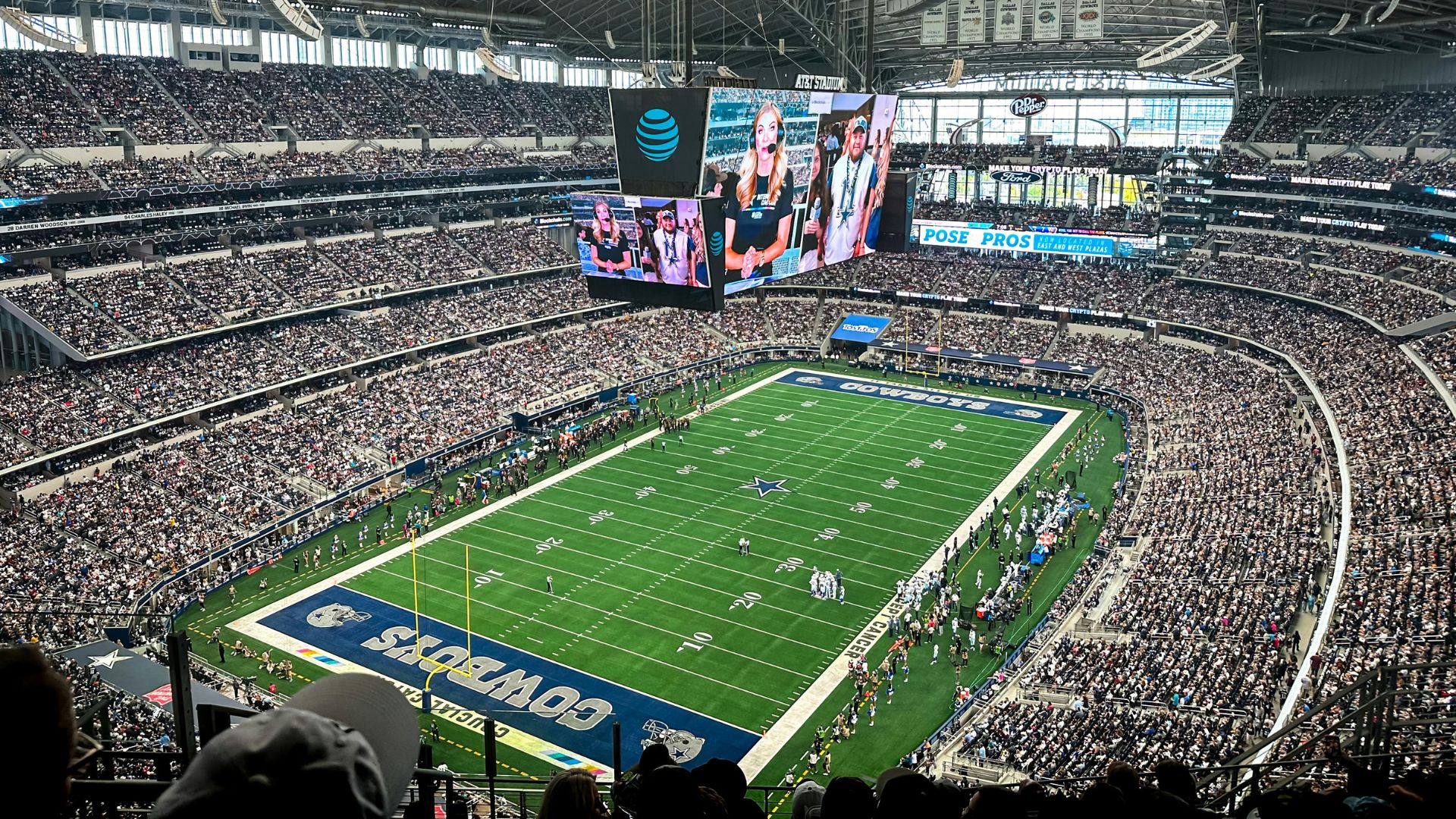 An overhead, wide-angle view of the packed AT&T Stadium football field and stands during an NFL game, with a large central video screen visible.