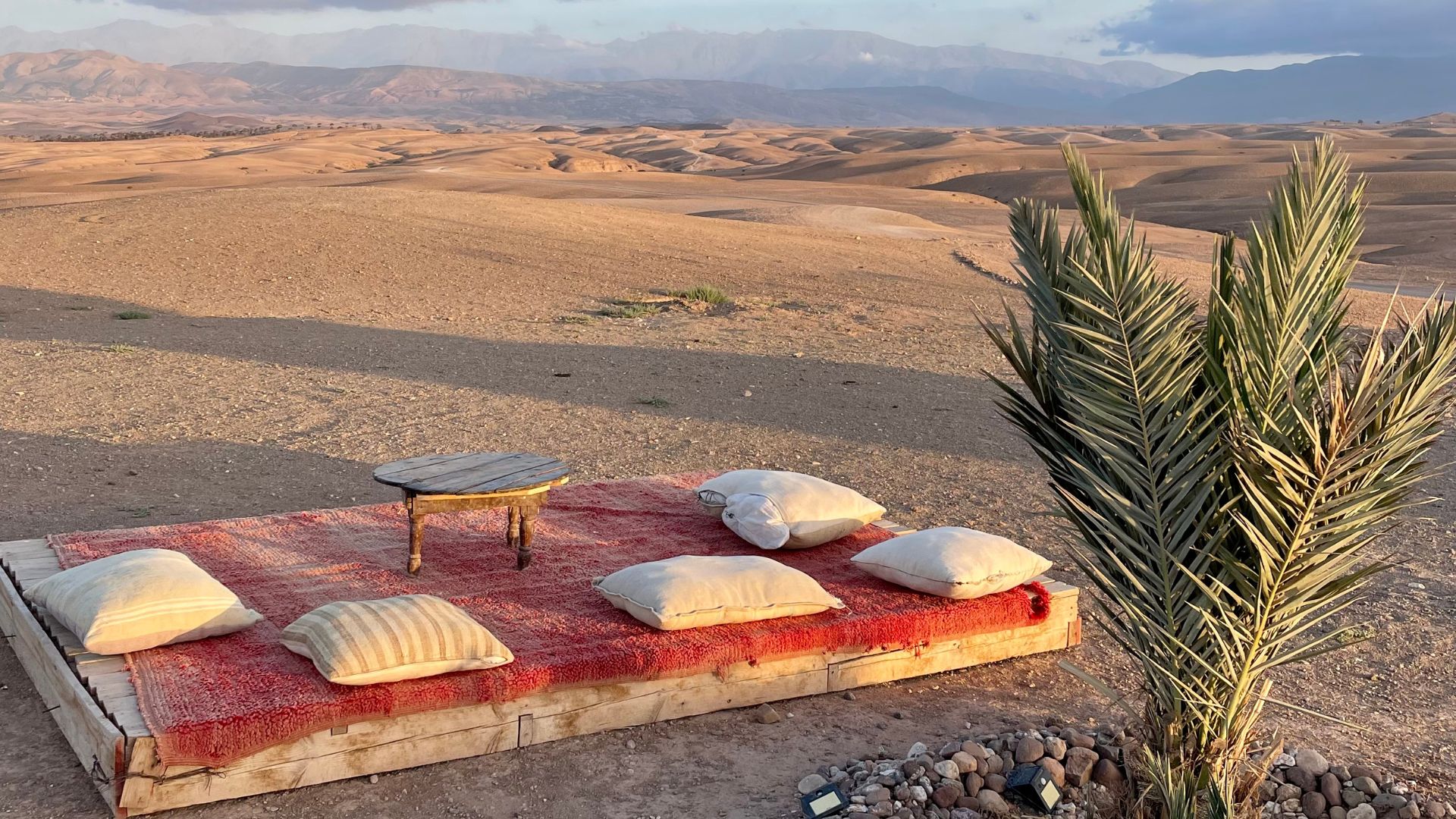 A comfortable outdoor seating area with cushions and a small table overlooking the rocky, arid landscape of the Agafay Desert in Morocco at sunset.