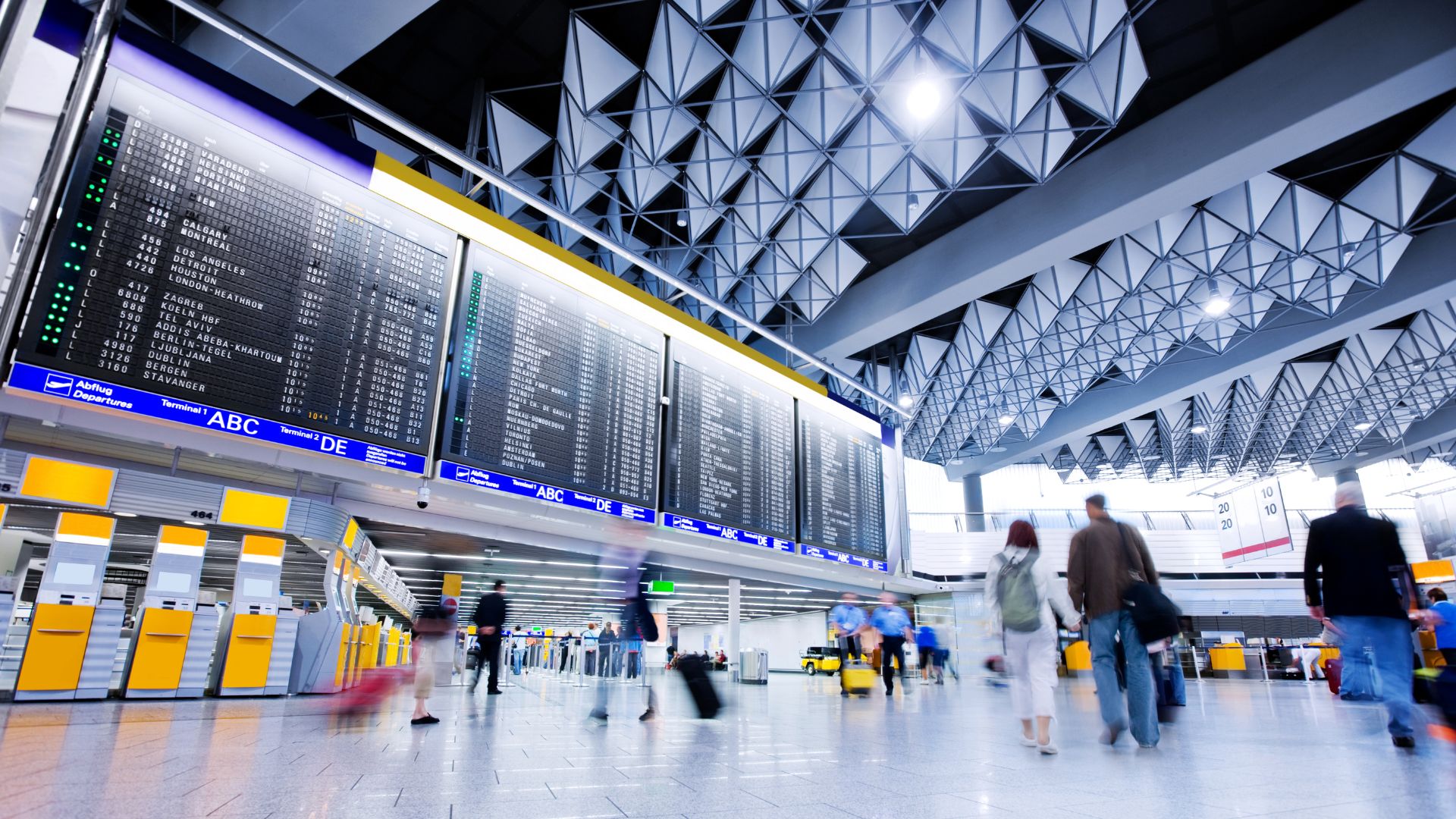 An interior wide shot of a busy airport terminal with a large flight departure board, high, modern triangular ceilings, and blurred figures of passengers walking with luggage.