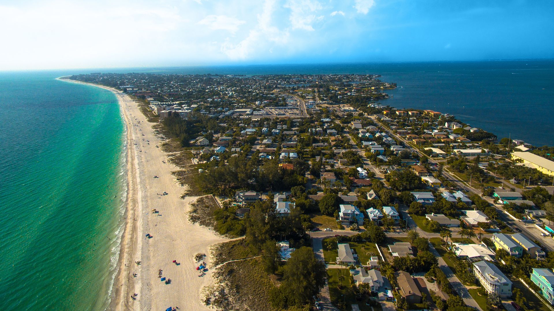 An aerial photograph of the northern tip of Anna Maria Island, Florida, showing a long, narrow stretch of land densely covered with houses and buildings between the bright turquoise waters of the Gulf of Mexico on one side and a bay on the other.