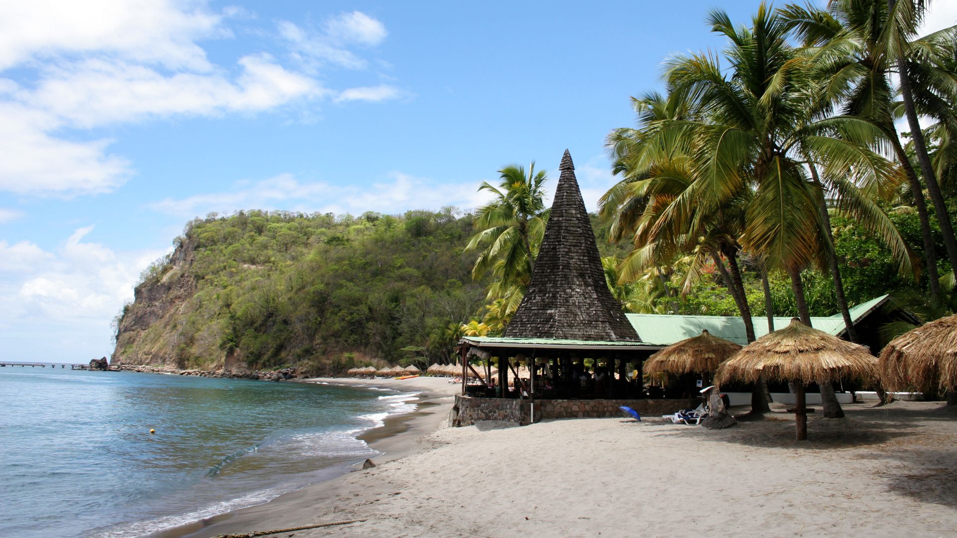 A scenic view of a sandy beach with palm trees and a thatched-roof structure, with a lush green headland extending into the sea under a blue sky with some clouds.
