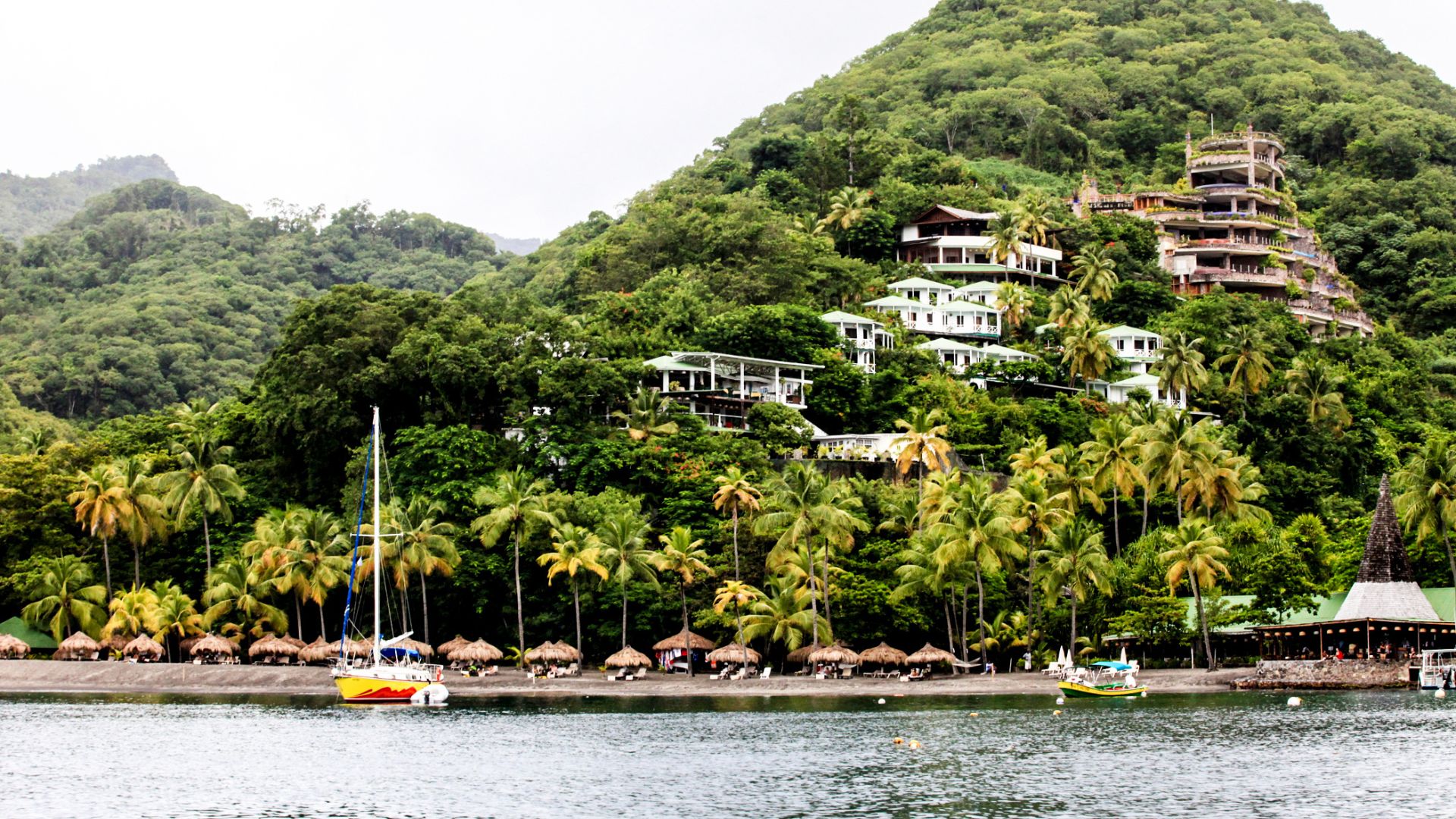 A tropical beach with palm trees and thatched huts, with a unique hillside resort built into the lush green mountain in the background.