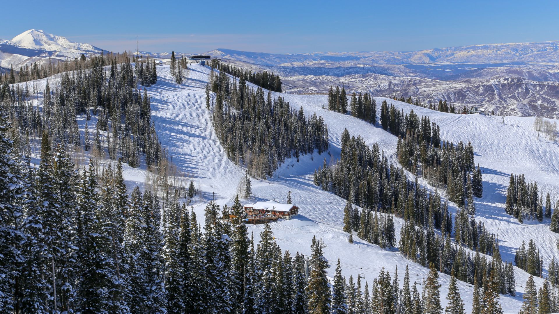 An aerial view of vast, snow-covered ski slopes lined with evergreen trees on a sunny mountain, with a large wooden lodge nestled on the side of a run and a panoramic view of distant snowy mountain ranges.