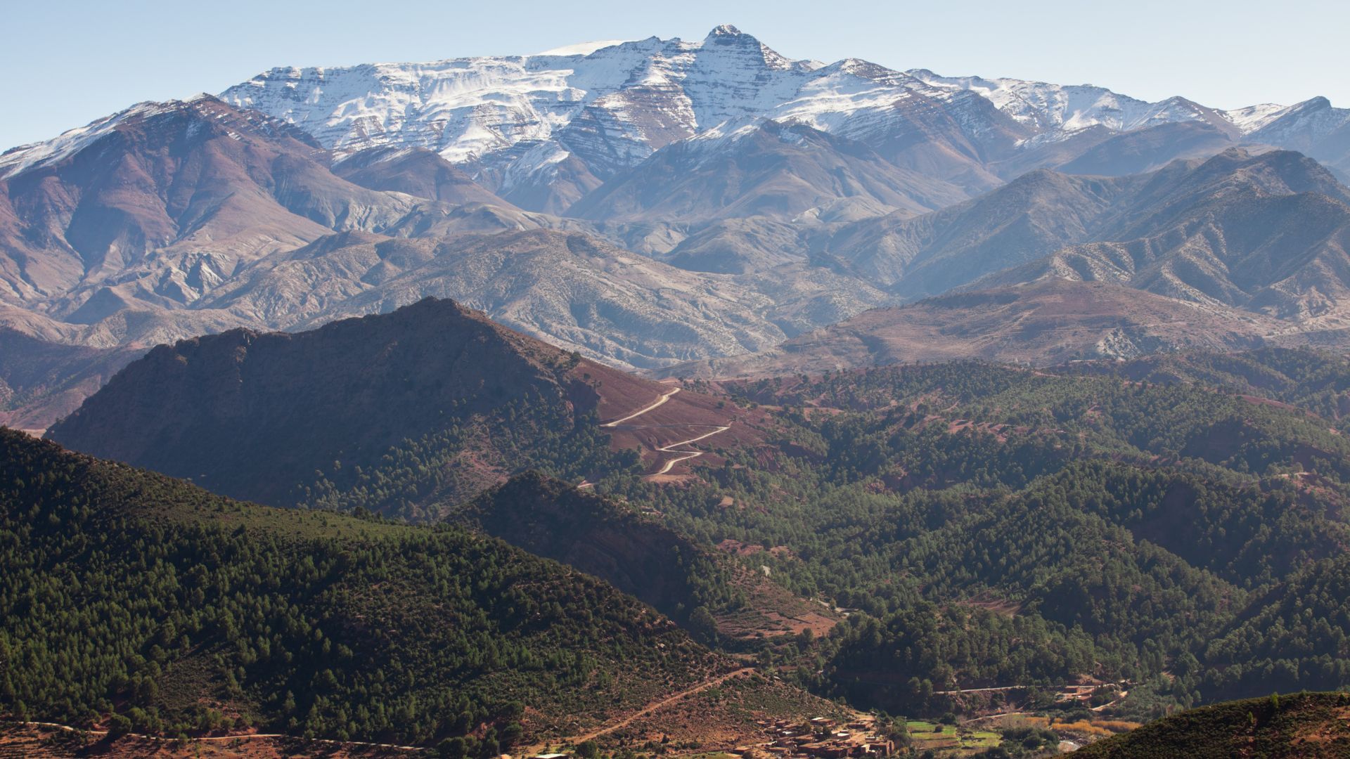 A panoramic view of rugged, tree-covered mountains with snow-capped peaks in the background.