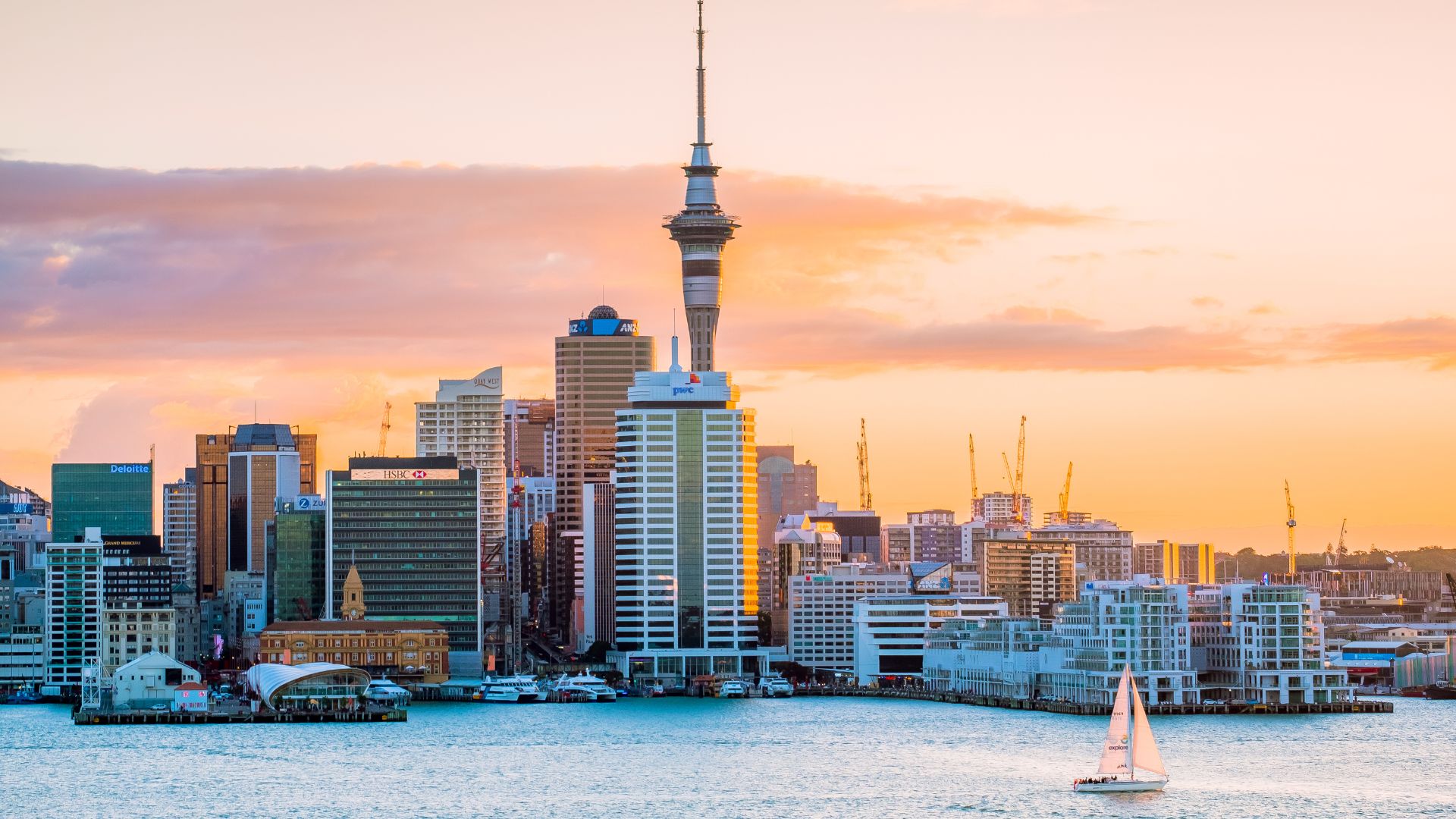 An aerial photograph of the Auckland, New Zealand, city skyline and harbor during sunset, featuring the tall, spire-like Sky Tower in the center, several skyscrapers, and a sailboat on the water in the foreground.