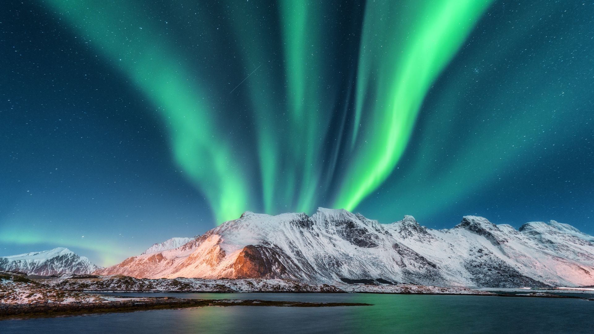 A night sky dominated by vibrant green, purple, and pink streaks of the Aurora Borealis reflecting on the calm water of a fjord, with large snow-covered mountains visible in the background along the distant shore.