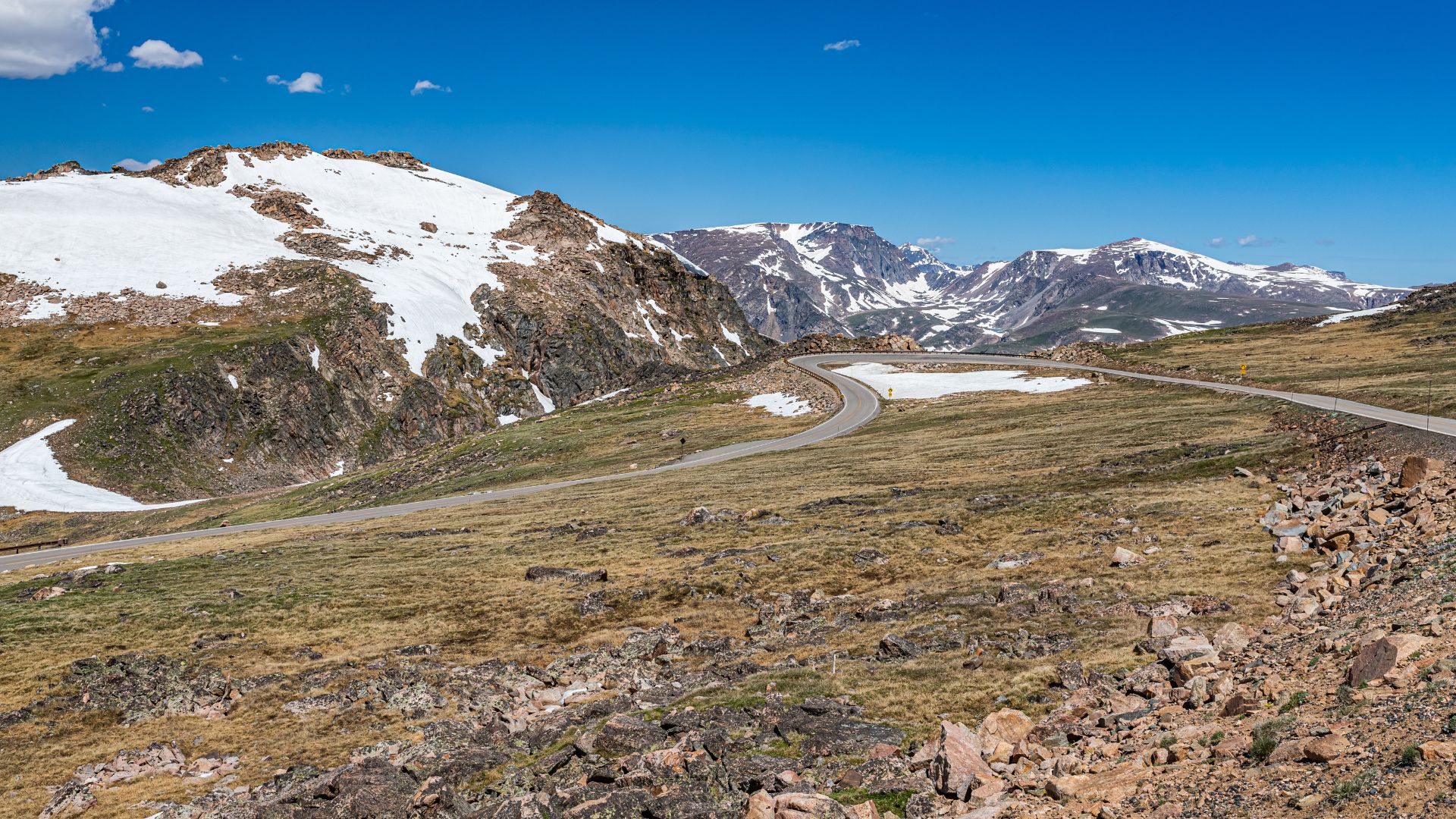 A two-lane mountain highway winds through a high-altitude, rocky landscape with snow patches and green grass, with rugged peaks visible in the distance under a blue sky.