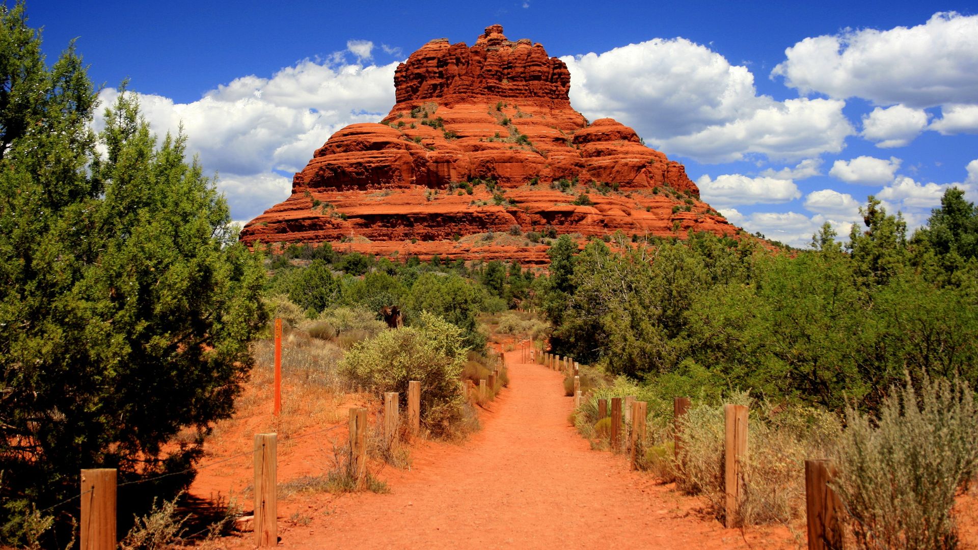 A prominent, layered red sandstone mountain known as Bell Rock towers over a desert landscape, with a dirt hiking trail marked by wooden posts leading towards it under a bright blue sky with white clouds.