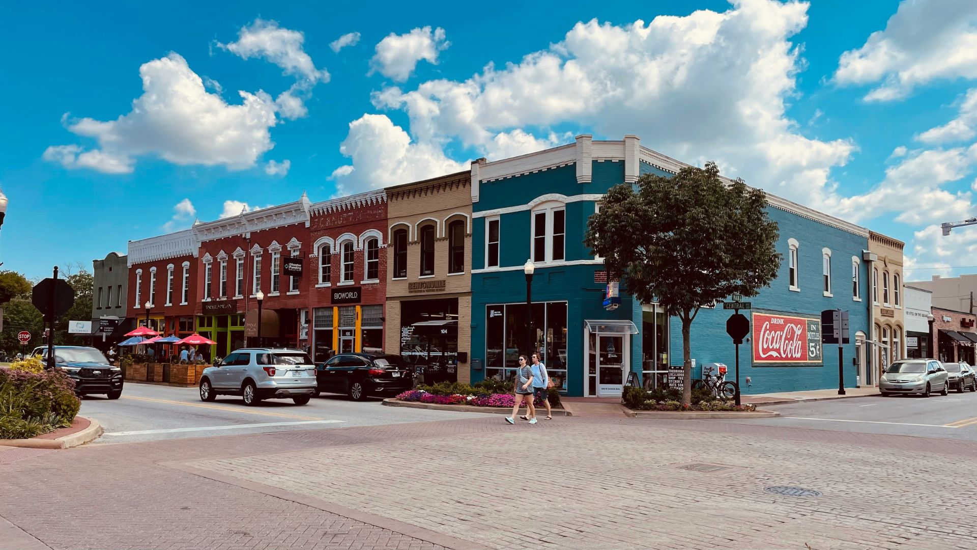 A daytime photo of a historic brick street corner lined with colorful, multi-story buildings, featuring several cars, pedestrians, and a prominent Coca-Cola sign on a teal building.