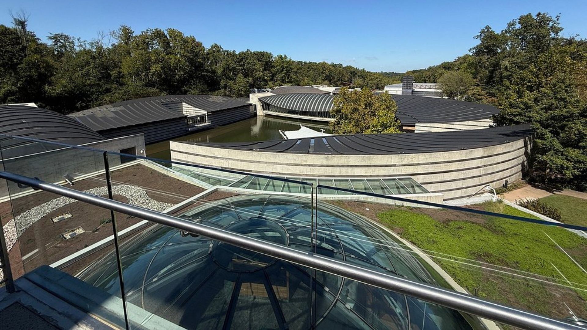 A complex of modern, curved buildings with dark, banded roofs and glass elements, nestled in a lush, green forest setting with water features.