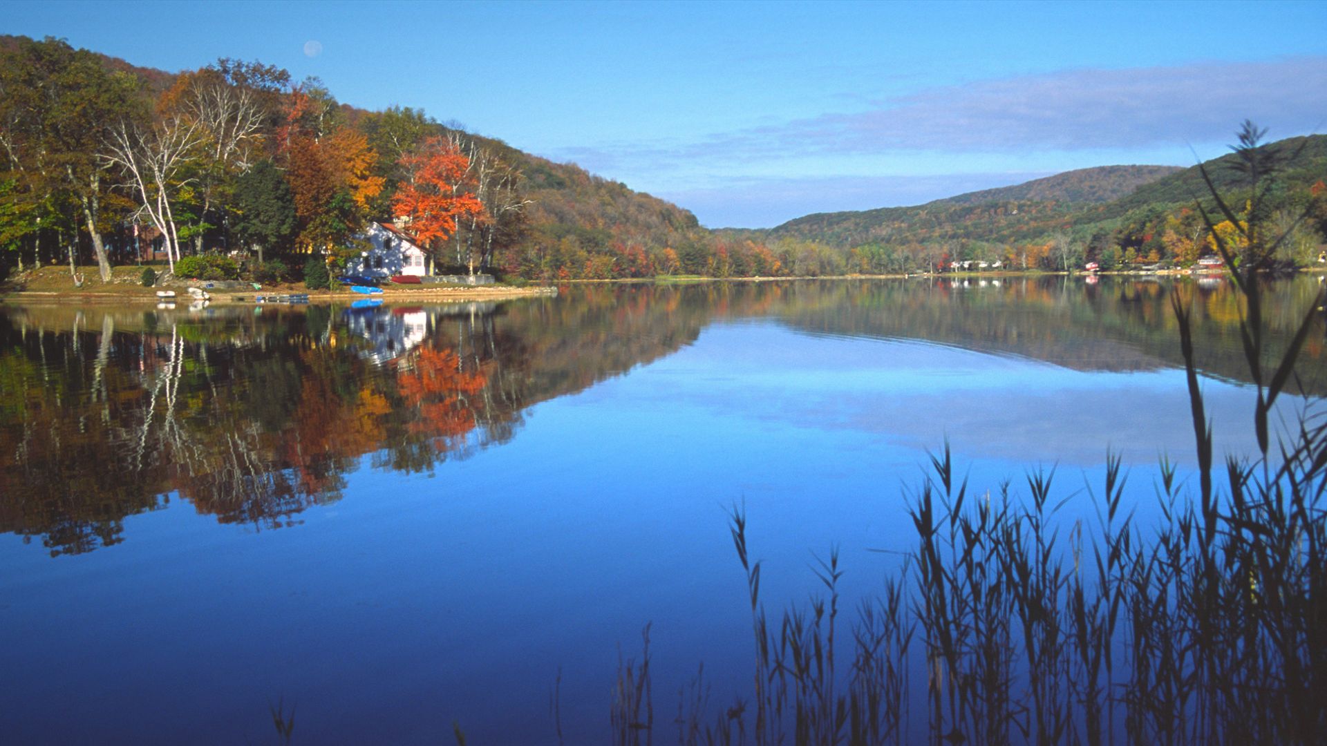 A serene lake reflects the vibrant orange, red, and yellow autumn foliage of the surrounding forested hills, with a white house visible on the left shoreline.