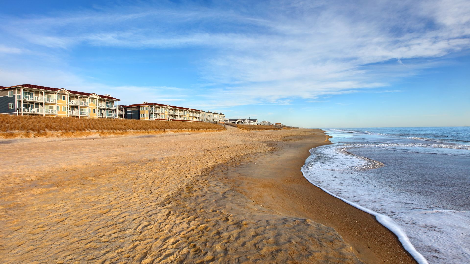 A wide, sandy beach with gentle ocean waves in the foreground, leading back to a row of yellow and white multi-story resort buildings nestled behind natural sand dunes under a blue sky with light clouds.