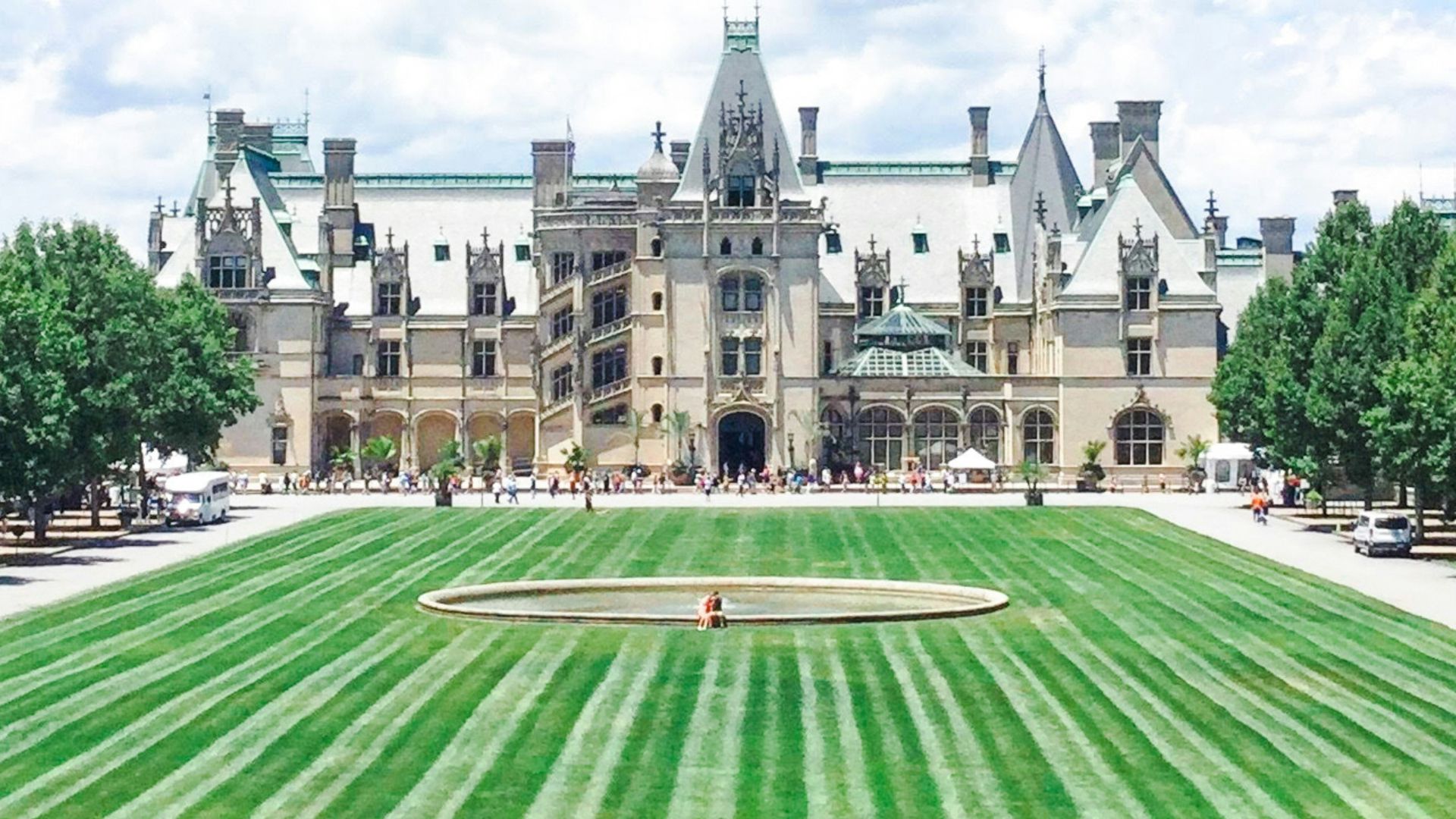 A bright, sunny day view of the magnificent, multi-story Biltmore Mansion with its ornate architecture and steepled turrets, featuring a wide, manicured green lawn in the foreground and a few people touring the grounds.