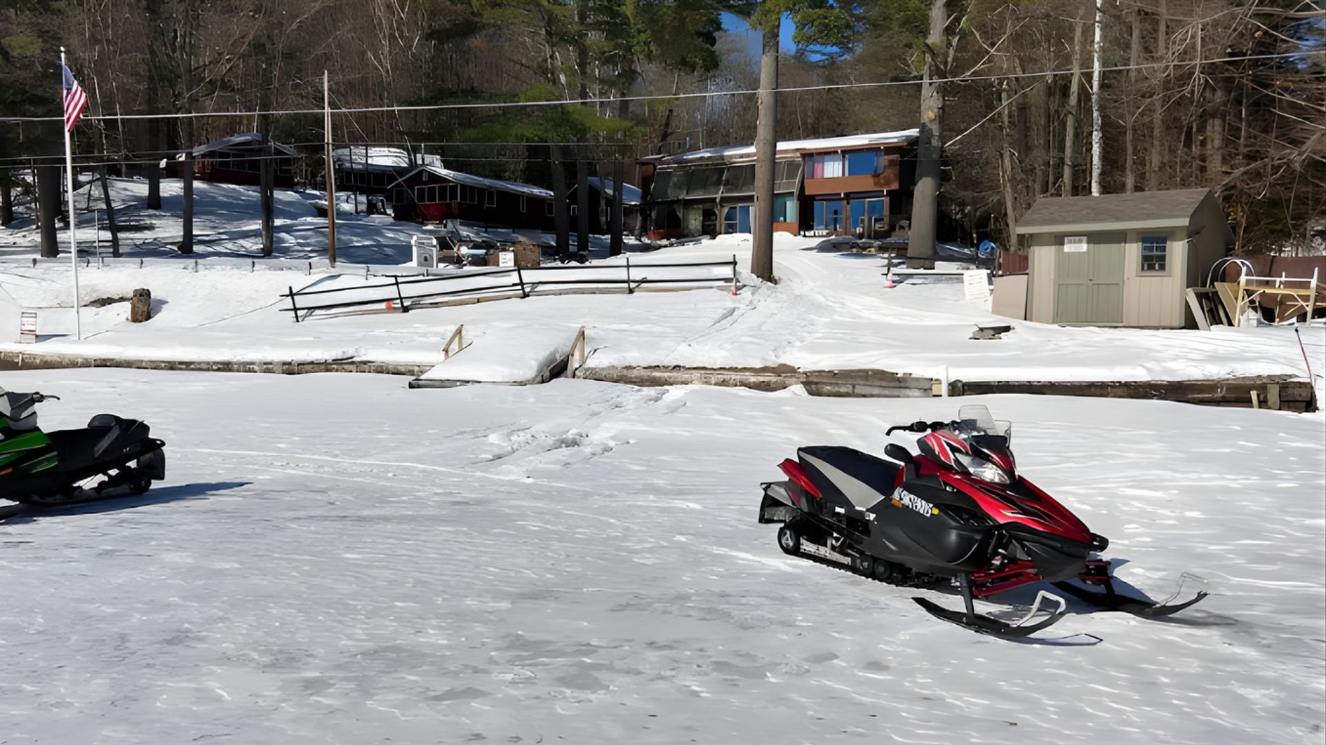 Two snowmobiles sit on a wide expanse of a frozen, snow-covered lake in front of several lake houses and a forested hill.