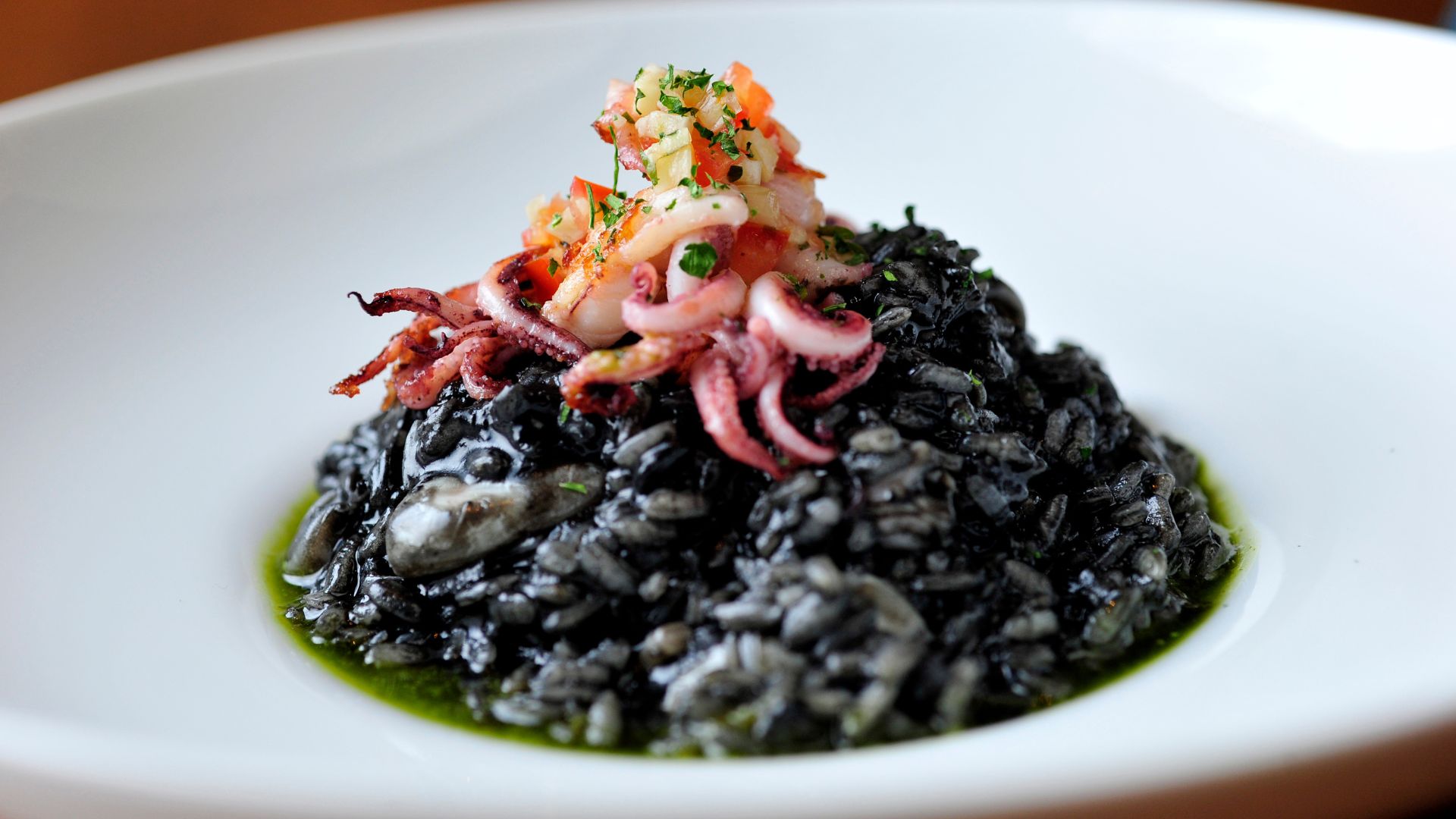 A close-up photograph of a plate of dark, glossy black risotto garnished with several cooked pink squid or octopus tentacles, diced red vegetables, and green herbs.