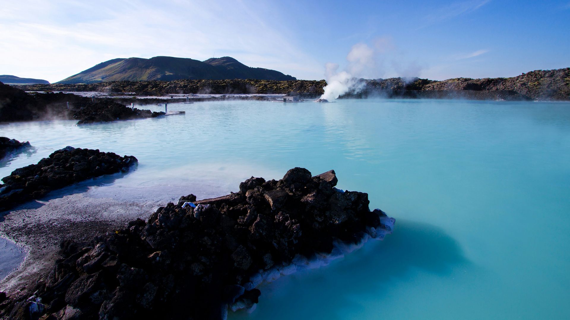 A vast, milky-blue geothermal lagoon with rising steam, surrounded by dark black lava rock formations and a hill in the distance under a blue sky.