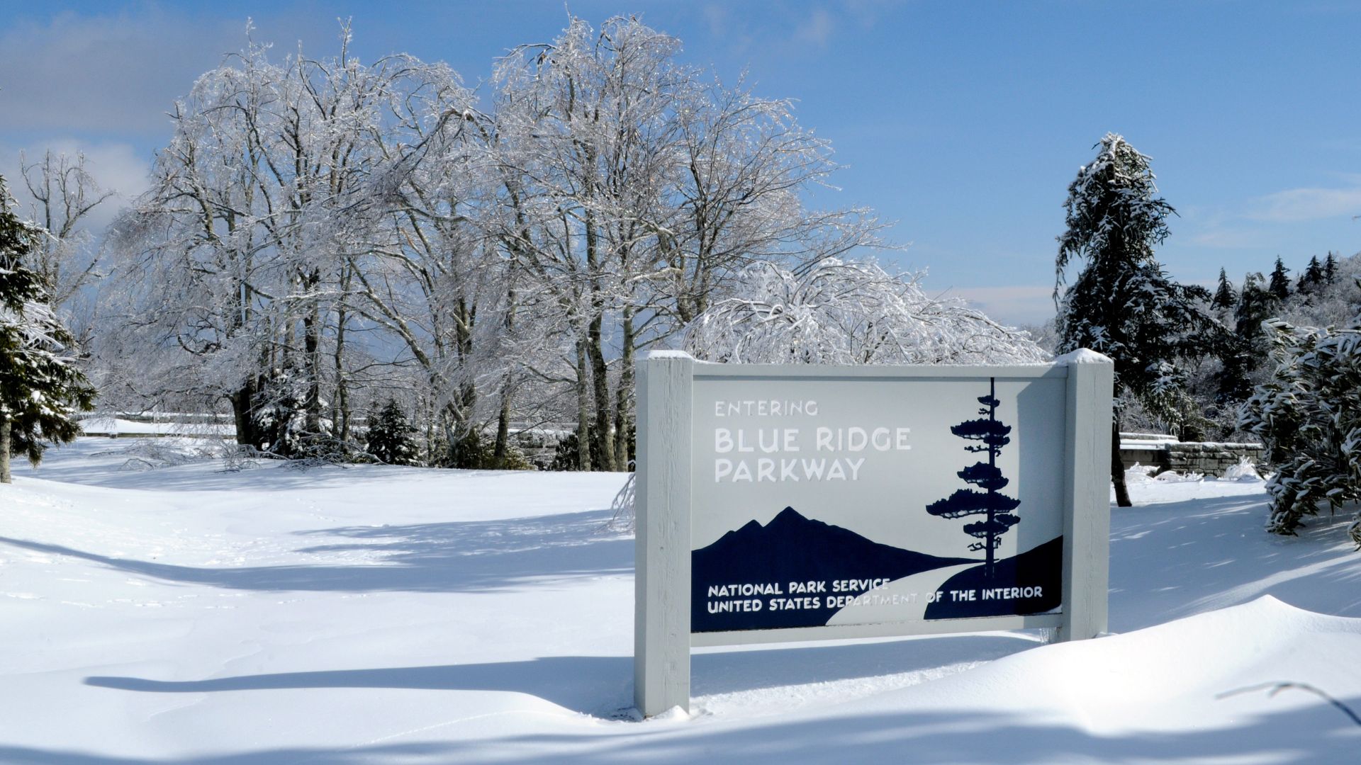 A white and blue national park sign for the "Blue Ridge Parkway" is surrounded by deep snow in a forest clearing on a bright winter day.