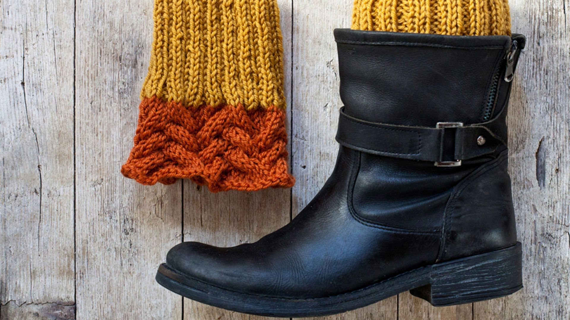 A pair of two-toned knitted boot cuffs (mustard yellow and orange cable knit) and a black leather ankle boot on a rustic wooden background.