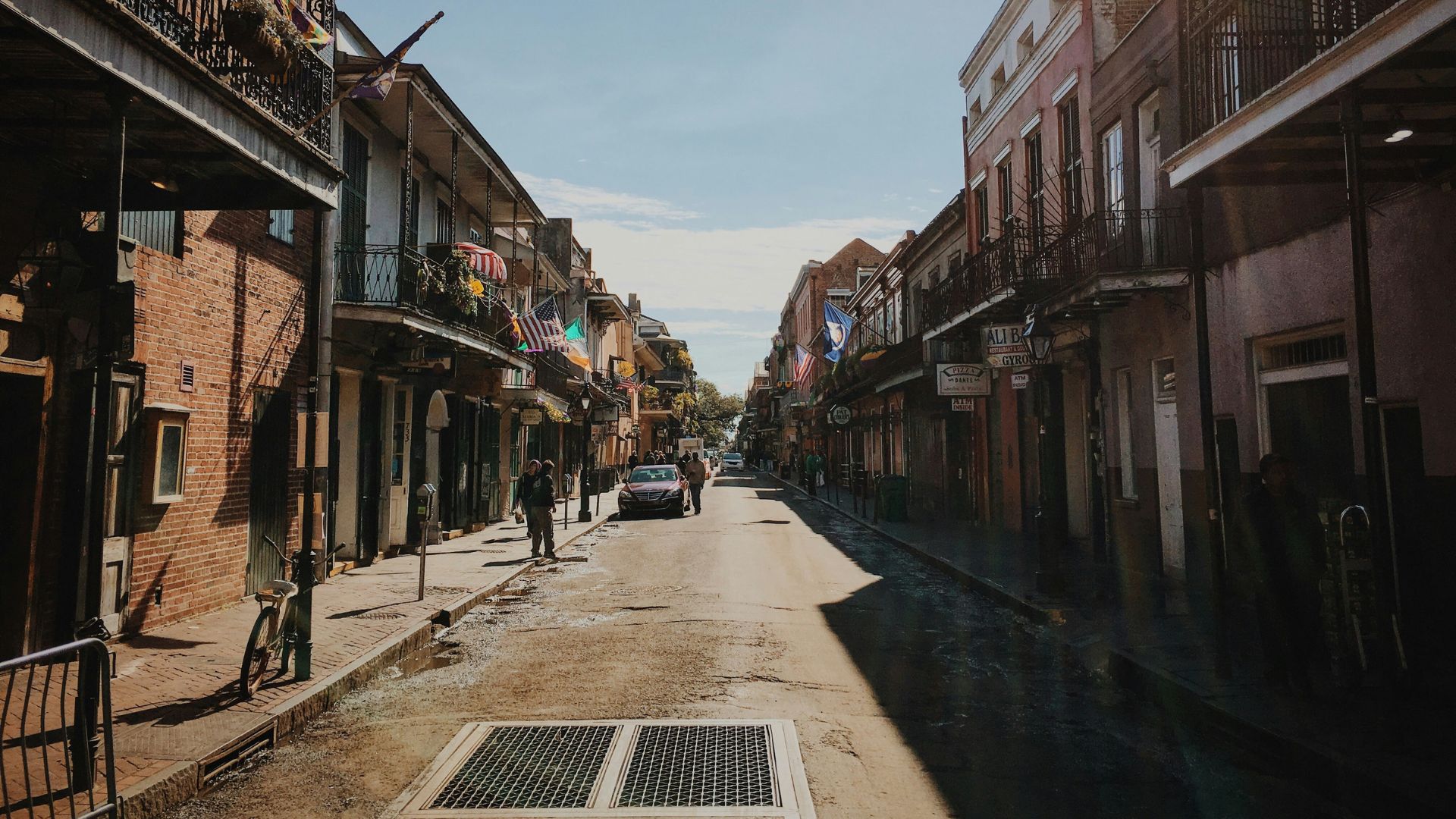 A sunny daytime view looking down the historic Bourbon Street in the French Quarter of New Orleans, featuring brick buildings with traditional iron balconies, a few people walking, and a car in the distance.