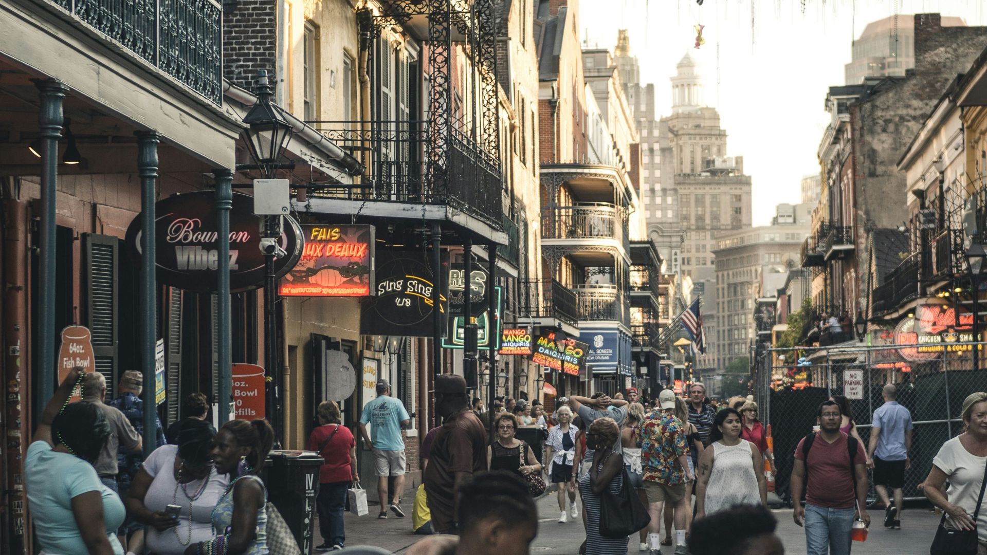 A daytime photo of a busy street in the French Quarter of New Orleans, lined with historic buildings featuring iron balconies. People are walking along the street, and two boys in the foreground are carrying buckets.