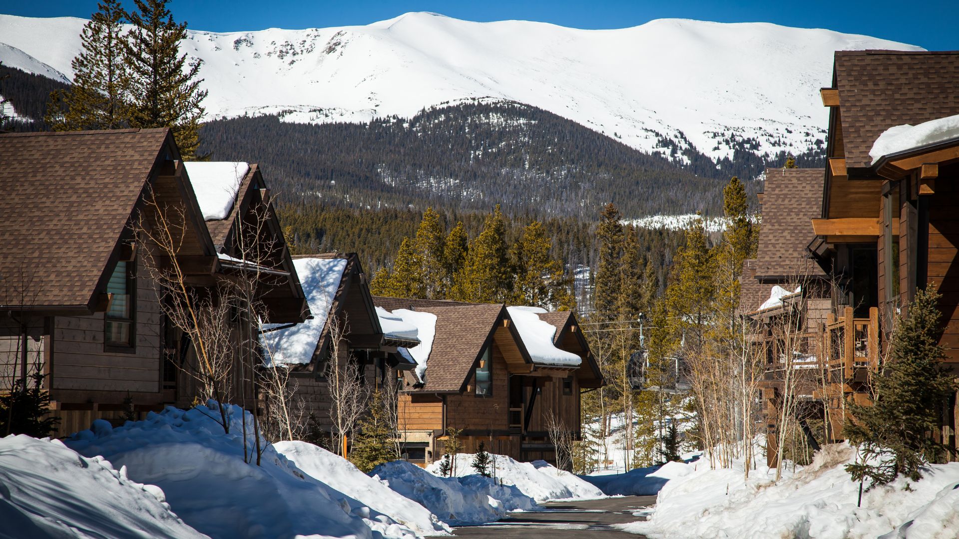 A view of rustic, multi-story wooden cabins and homes covered in snow, located in a resort community with a dense evergreen forest and large, snow-capped mountains in the background in Breckenridge, Colorado.