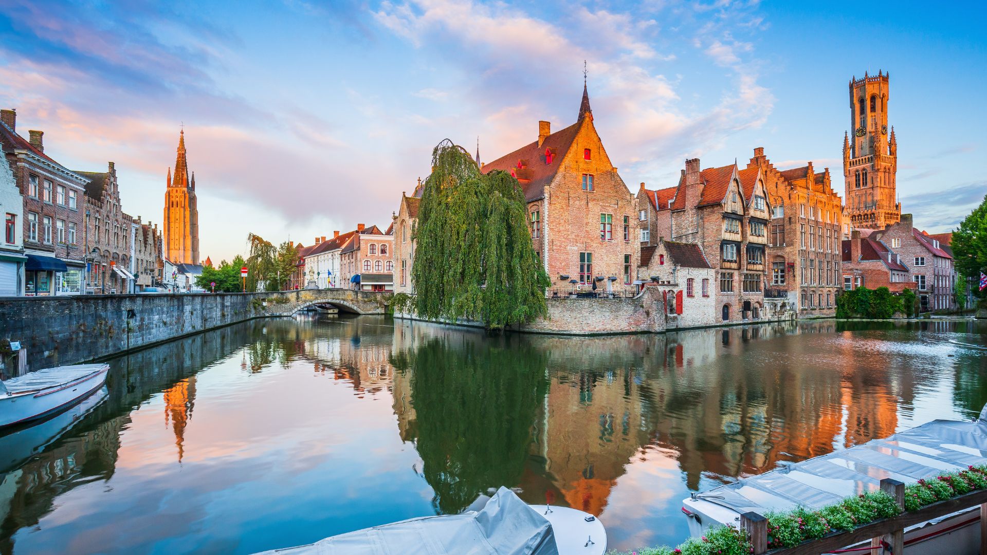 A beautiful, sunrise photograph of the historic canals in Bruges, Belgium, with reflections of medieval buildings, the tall spire of the Church of Our Lady, and the square Belfry tower visible in the distance.