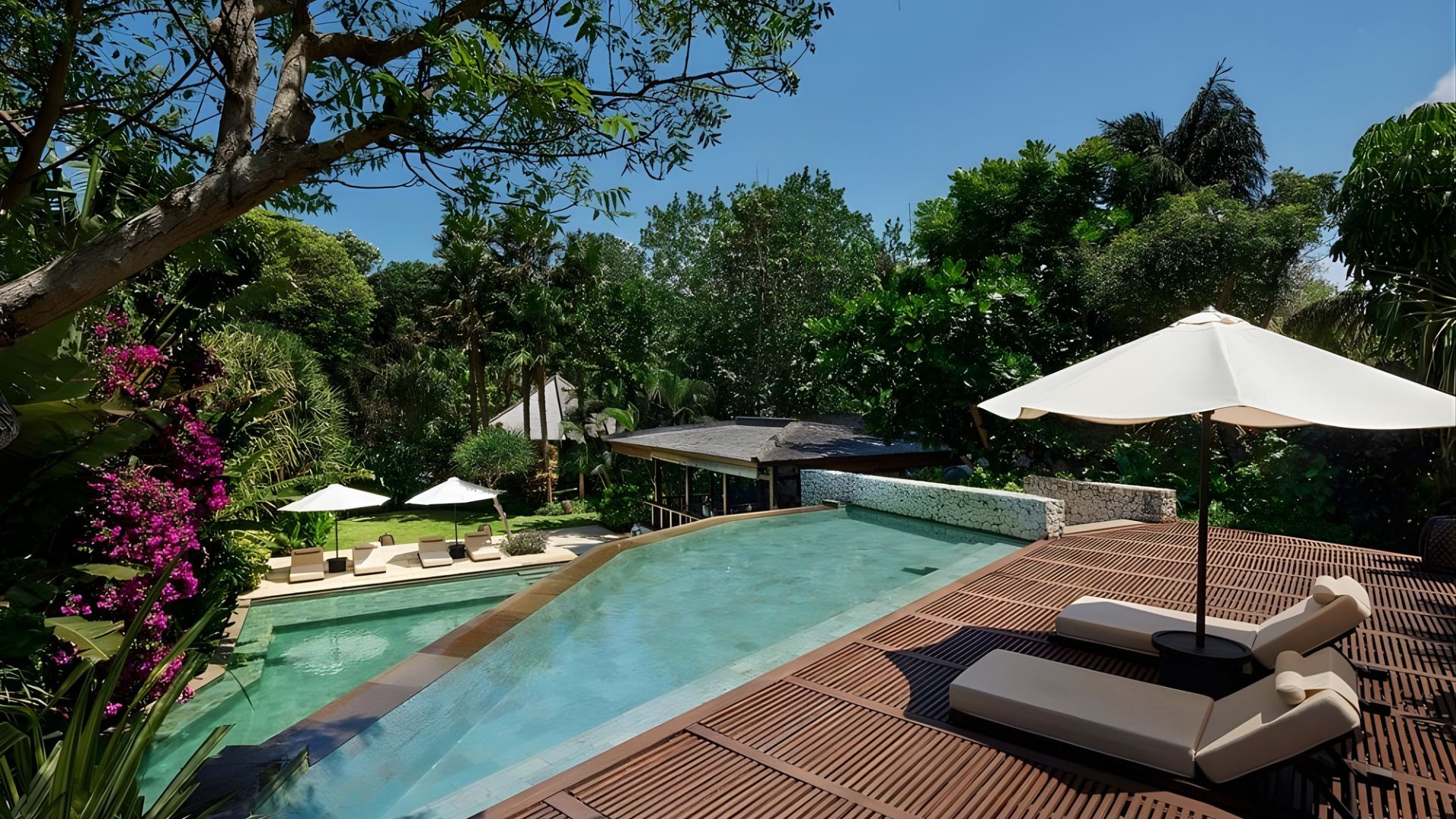 A view of a tiered swimming pool with a wooden deck and lounge chairs, set amidst lush green tropical gardens at a luxury resort in Bali.