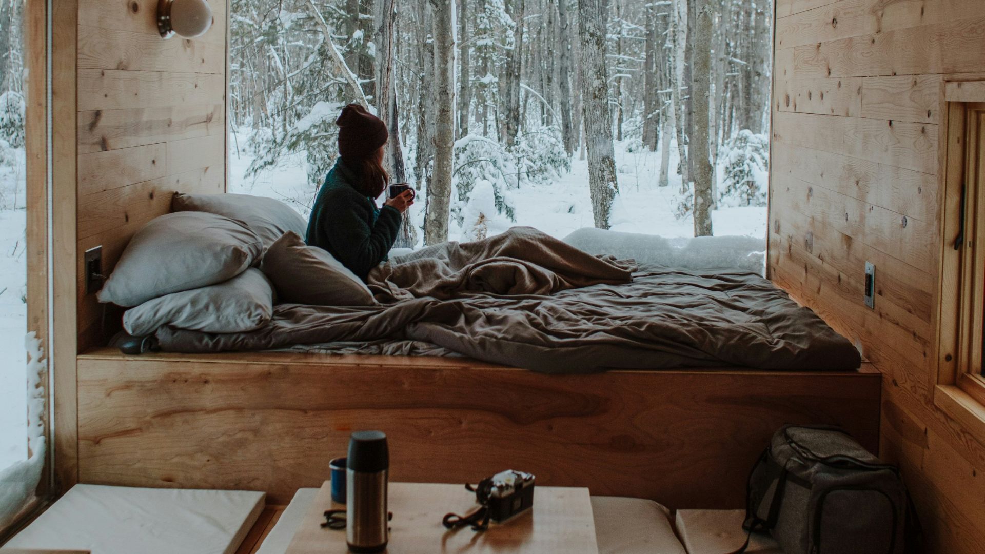 A person sits on a bed inside a small, wood-paneled cabin, looking out a large window at a snowy forest.