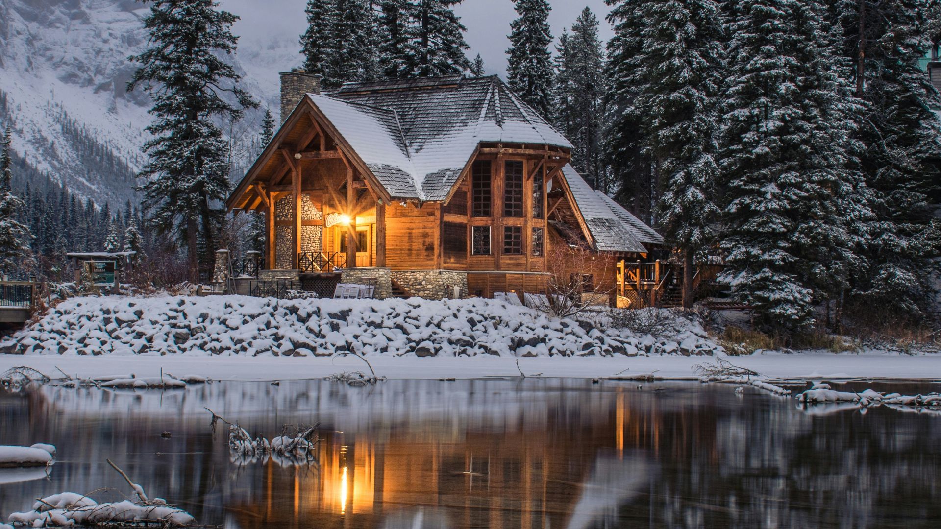 A large, warm, log cabin sits by a snowy lake at dusk, reflecting in the water amidst tall pine trees.