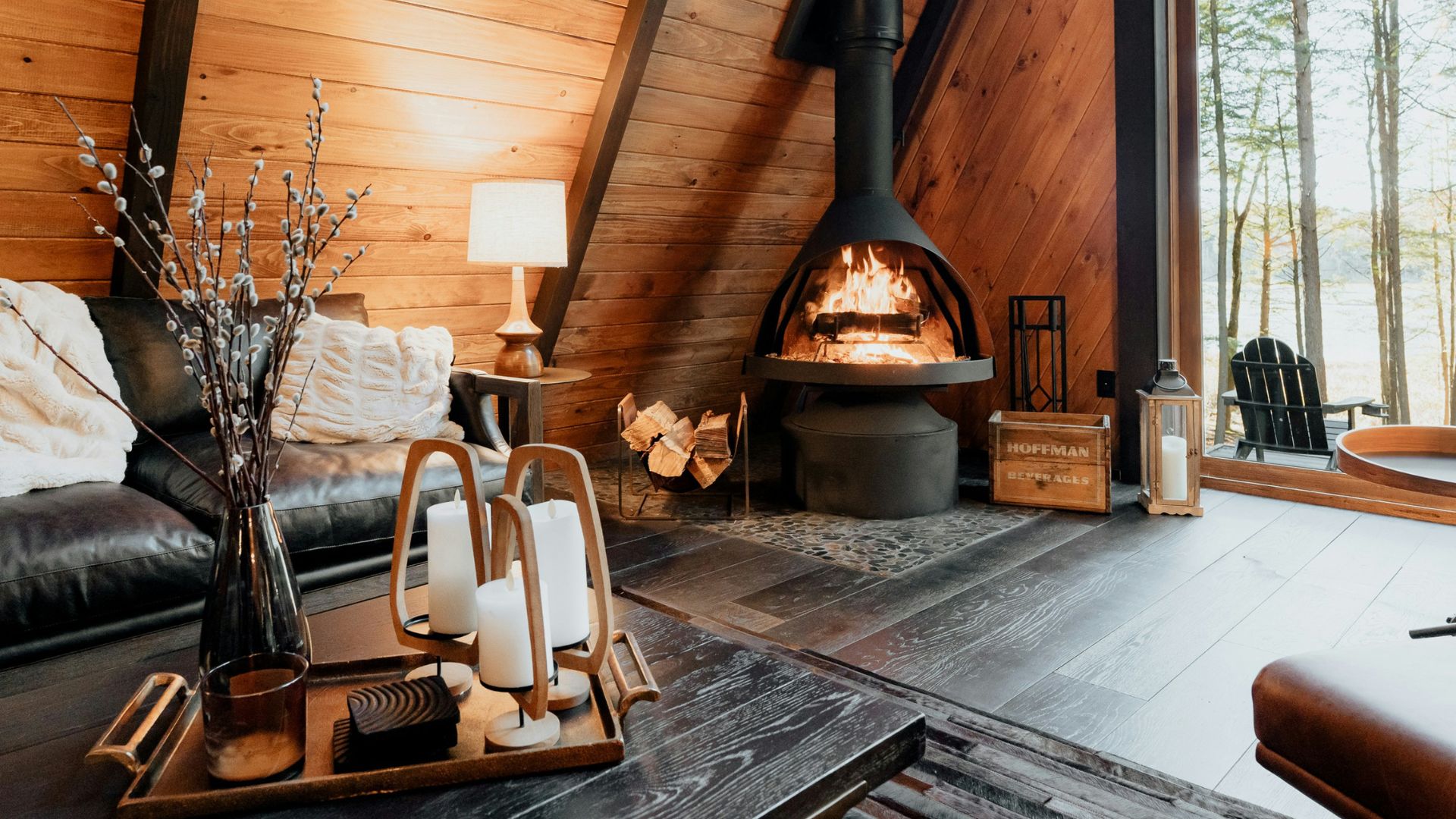 The cozy, wood-paneled interior of an A-frame cabin featuring a modern black wood-burning stove, a leather sofa, and a large window looking out onto trees.