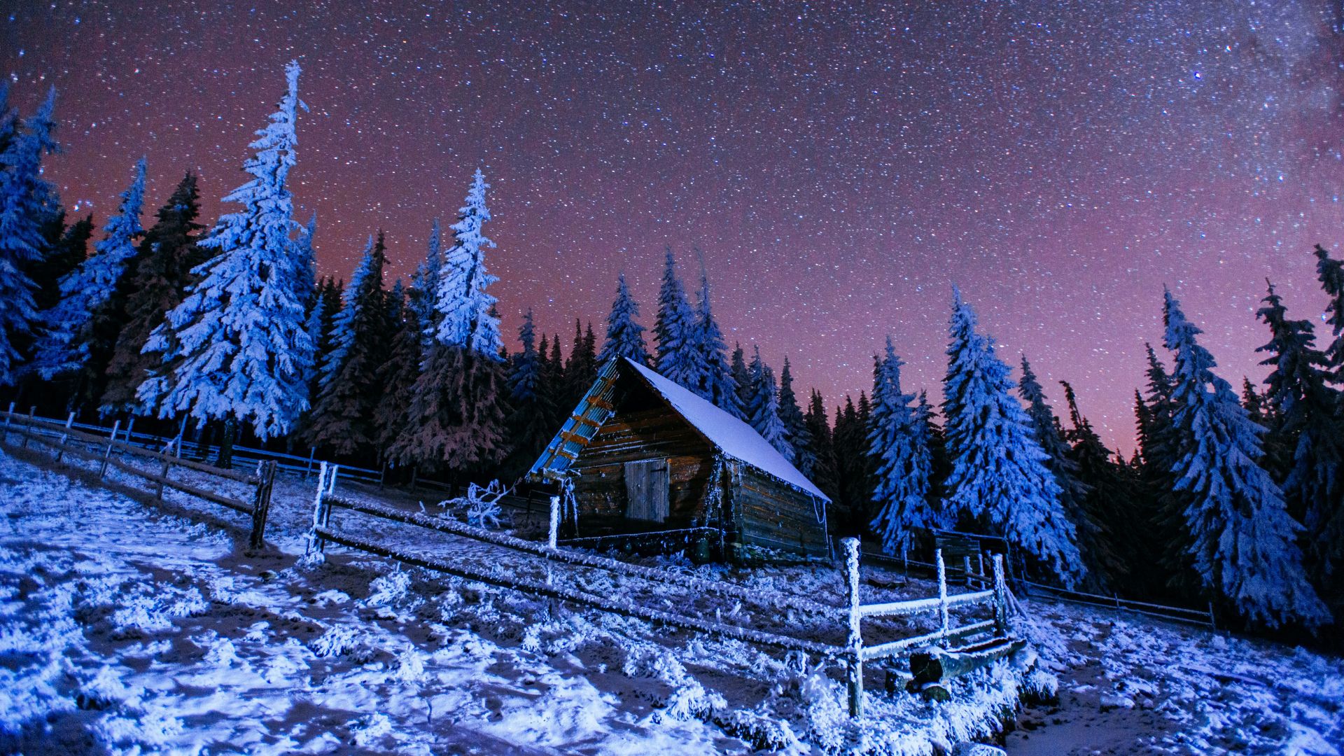 A small, rustic wooden cabin nestled in a snowy forest at night, under a star-filled, purple and pink sky.