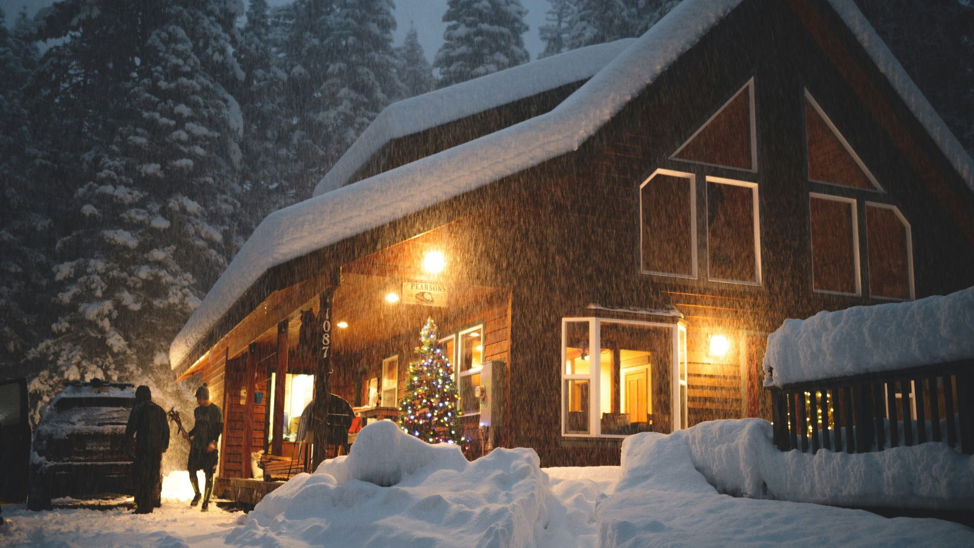 A warm, lit-up wooden cabin covered in deep snow during a snowy night, with two people and a car parked outside.