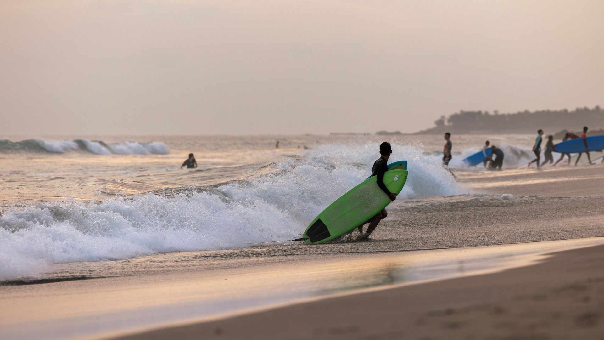 A person with a green surfboard walking into the waves at Canggu Beach during sunset, with other people in the background.