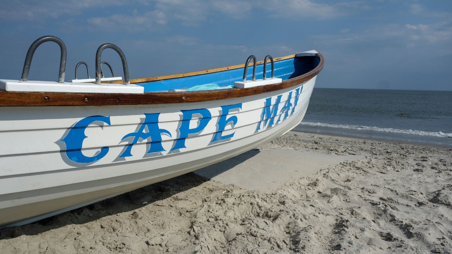 A white and blue wooden Cape May lifeguard rescue boat resting on a wide, sandy beach, with gentle ocean waves in the background under a blue sky.