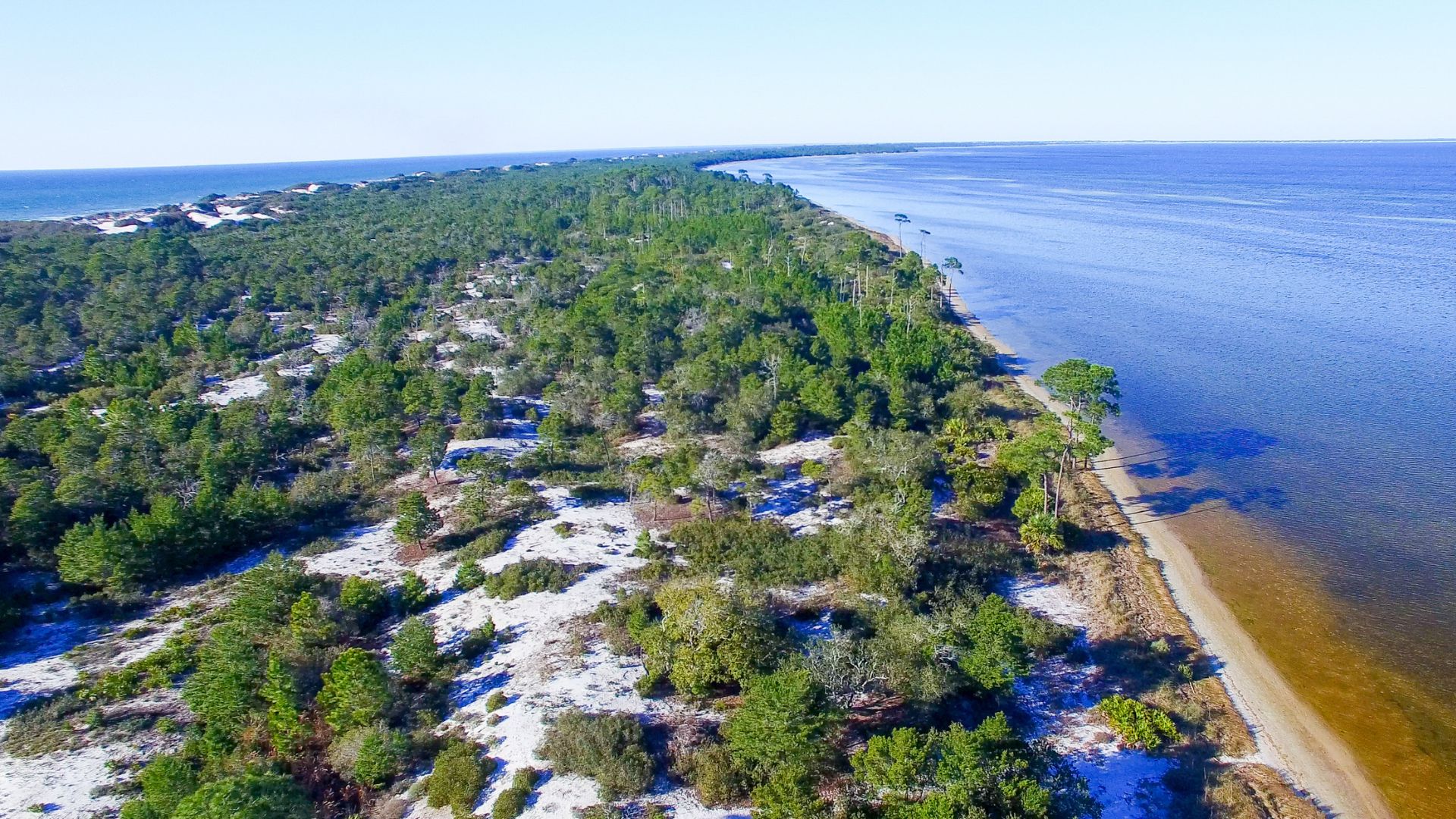 An aerial view of a long, narrow, tree-covered peninsula with white sand on both the bay side (right) and the open water side (left) on a clear, sunny day.