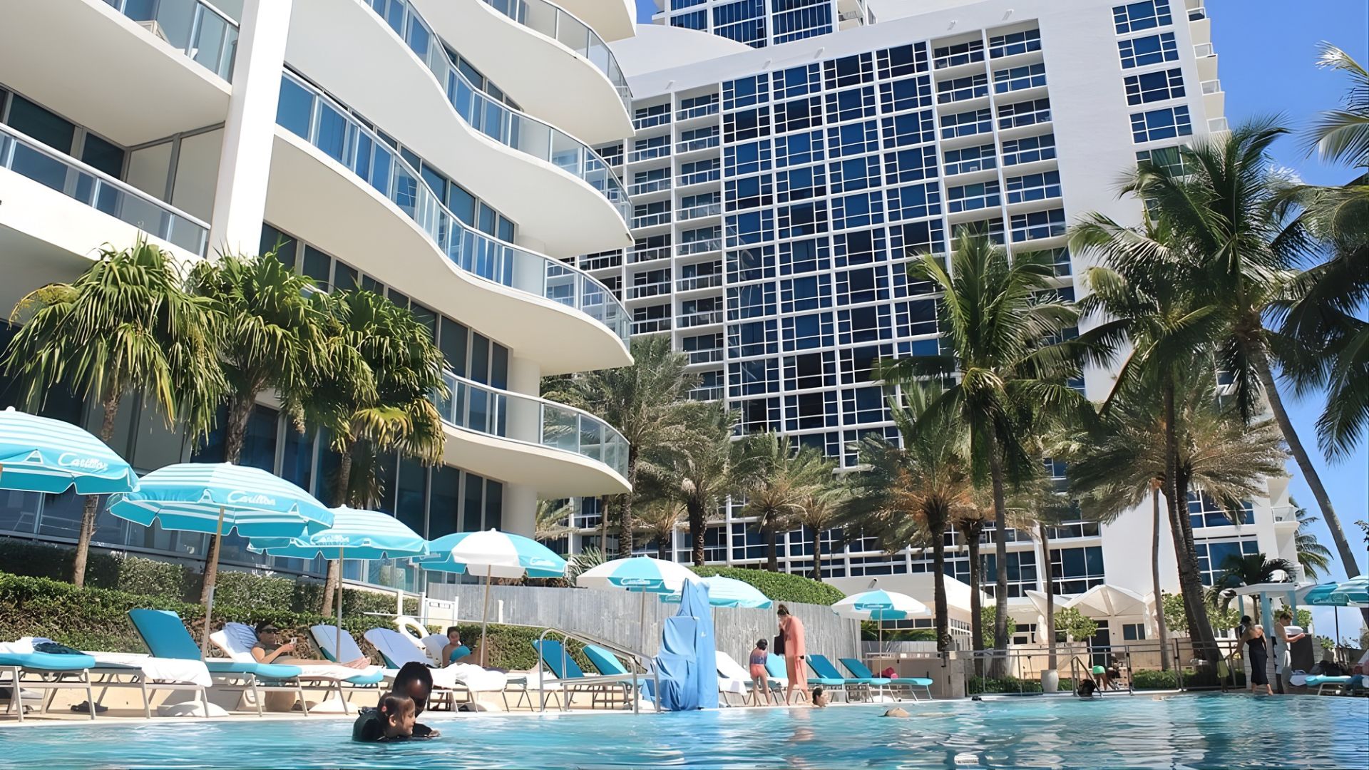 An outdoor view of two high-rise hotel or condominium buildings with a large swimming pool and palm trees in the foreground.