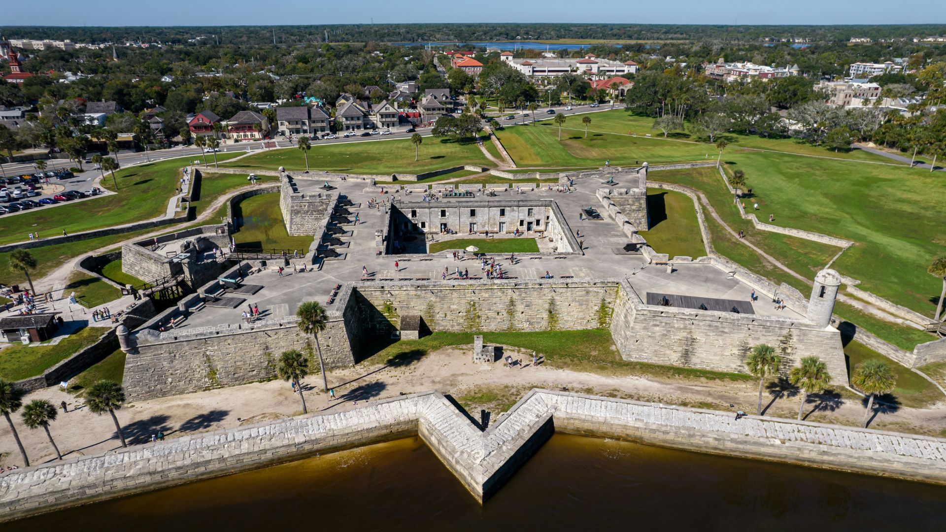 An aerial view of the star-shaped Castillo de San Marcos fort surrounded by a dry moat and green grass, with a city visible in the background.