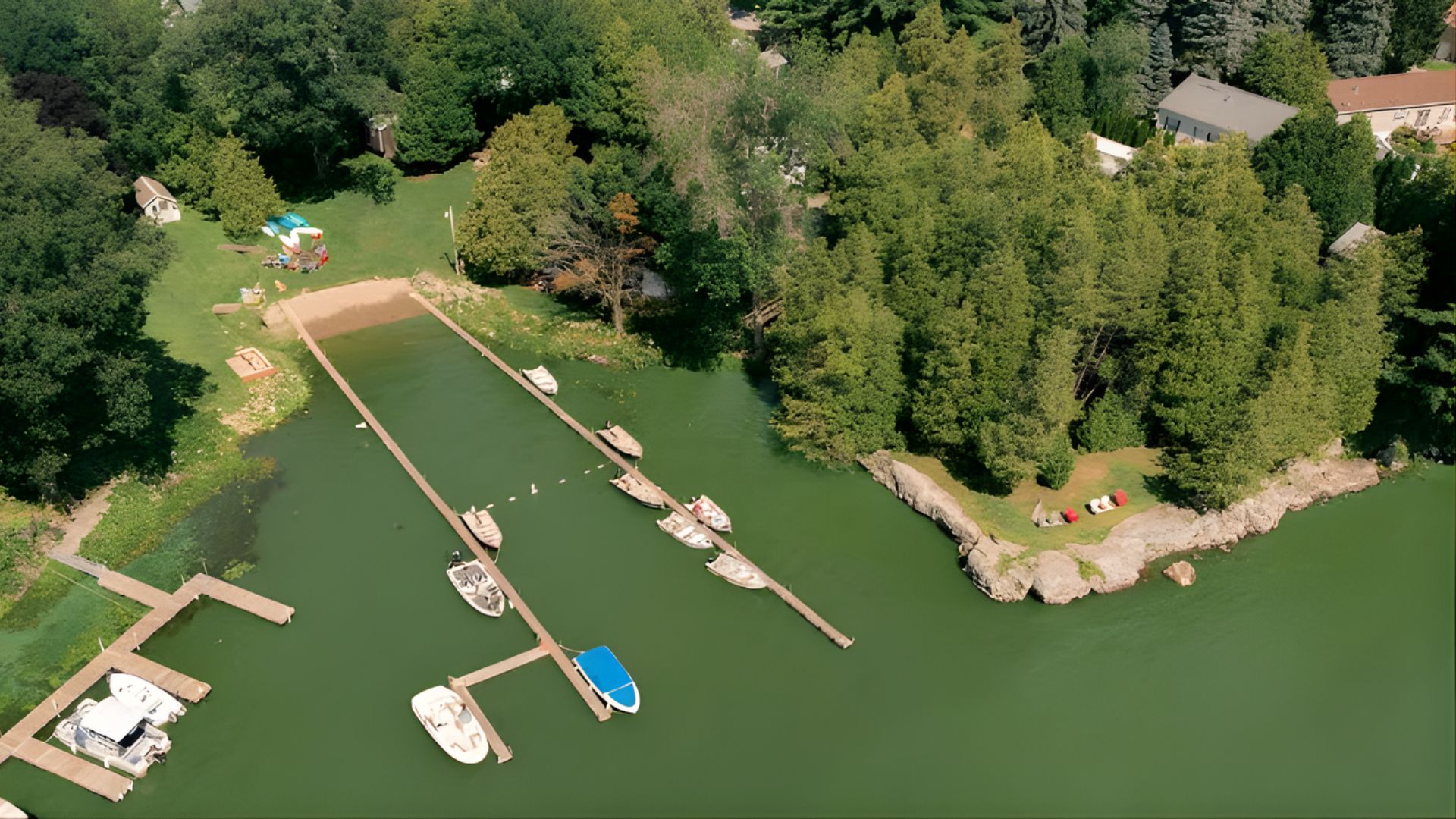 An aerial view of a lakefront resort with several long wooden docks extending into a calm, green-tinted lake, with a small sandy beach area on the shore surrounded by lush green trees.
