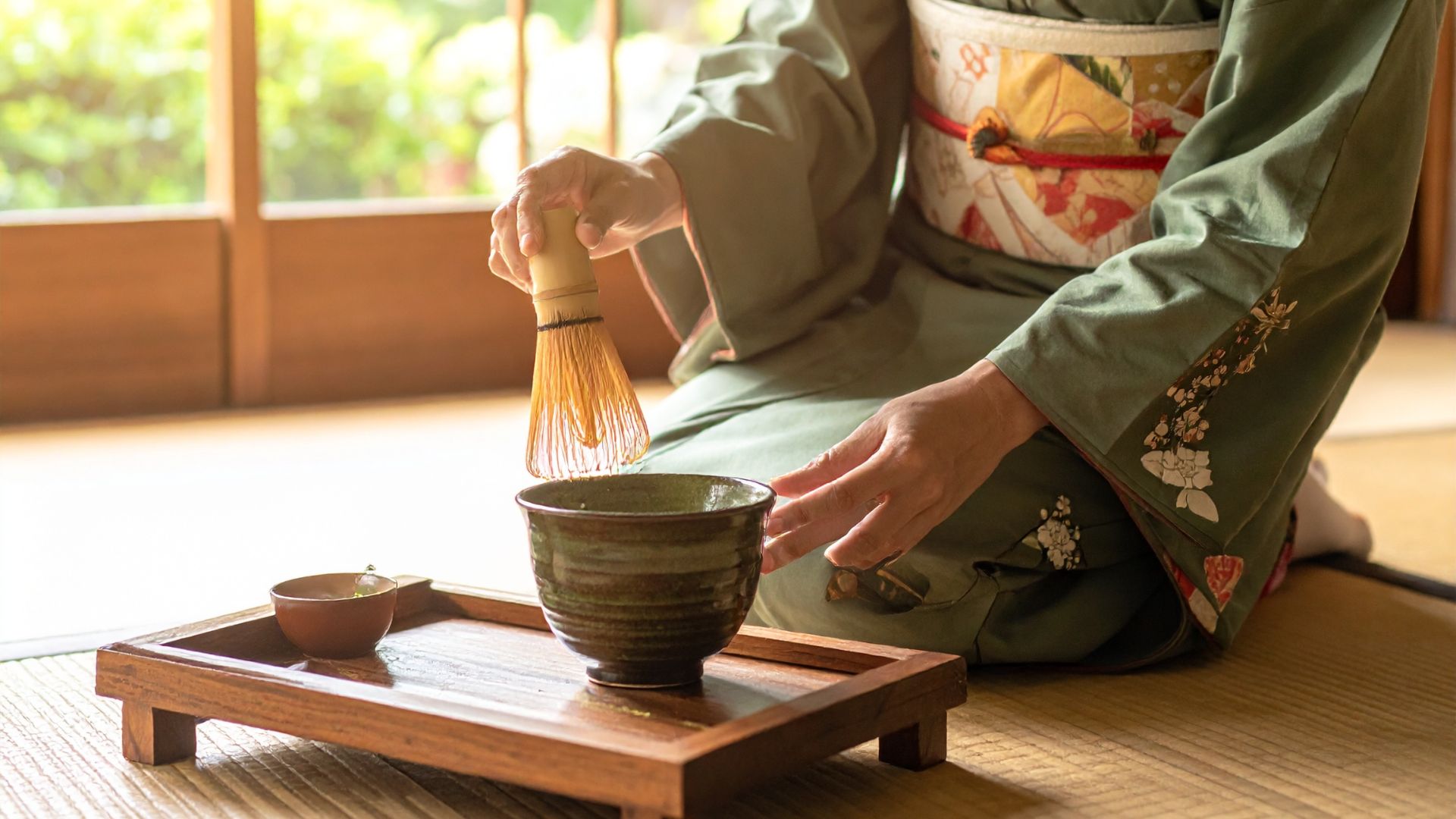 A person wearing a traditional green kimono kneels on a tatami mat, using a bamboo whisk to prepare matcha tea in a rustic ceramic bowl during a Japanese tea ceremony.