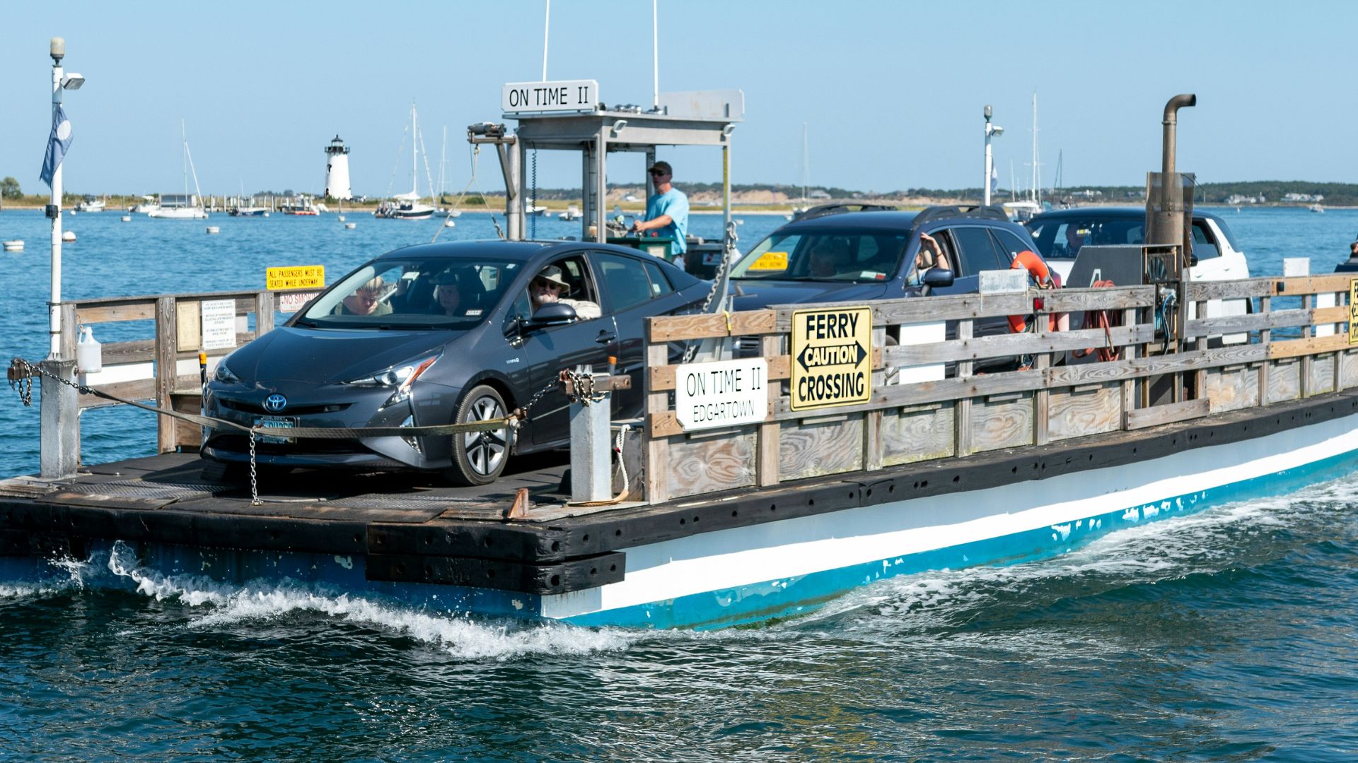 A small, open-air ferry carrying two cars across a calm harbor on a sunny day, with other boats and a lighthouse visible in the background.