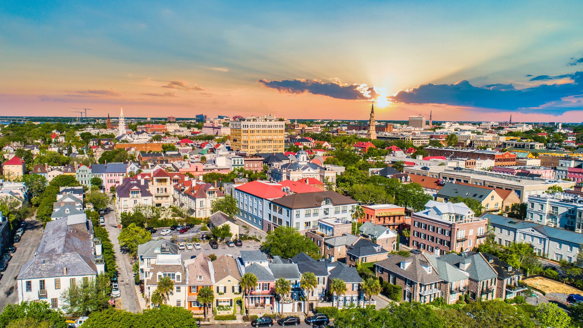 An elevated photo of the historic Charleston, South Carolina, skyline at sunset, showing many traditional buildings with colorful roofs and prominent church steeples, surrounded by lush green trees under a vibrant sky with sunbeams breaking through the clouds.
