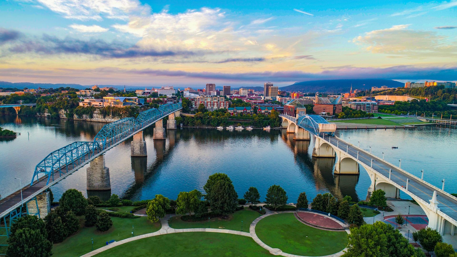 An aerial view of the Chattanooga, Tennessee skyline at sunset, featuring a blue pedestrian truss bridge and a concrete arch bridge spanning the wide Tennessee River.