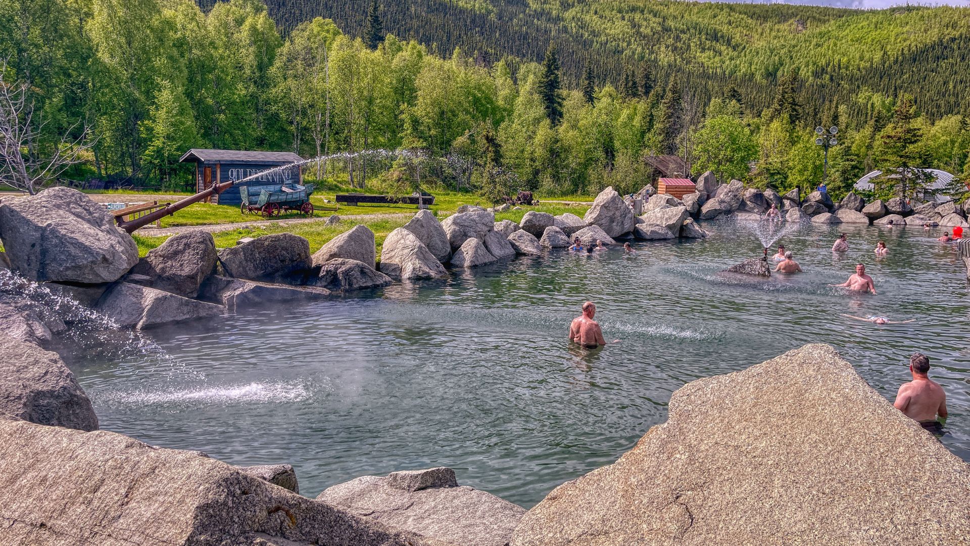 People soaking in an outdoor, rock-lined hot springs pool surrounded by green trees and a small cabin in the background.