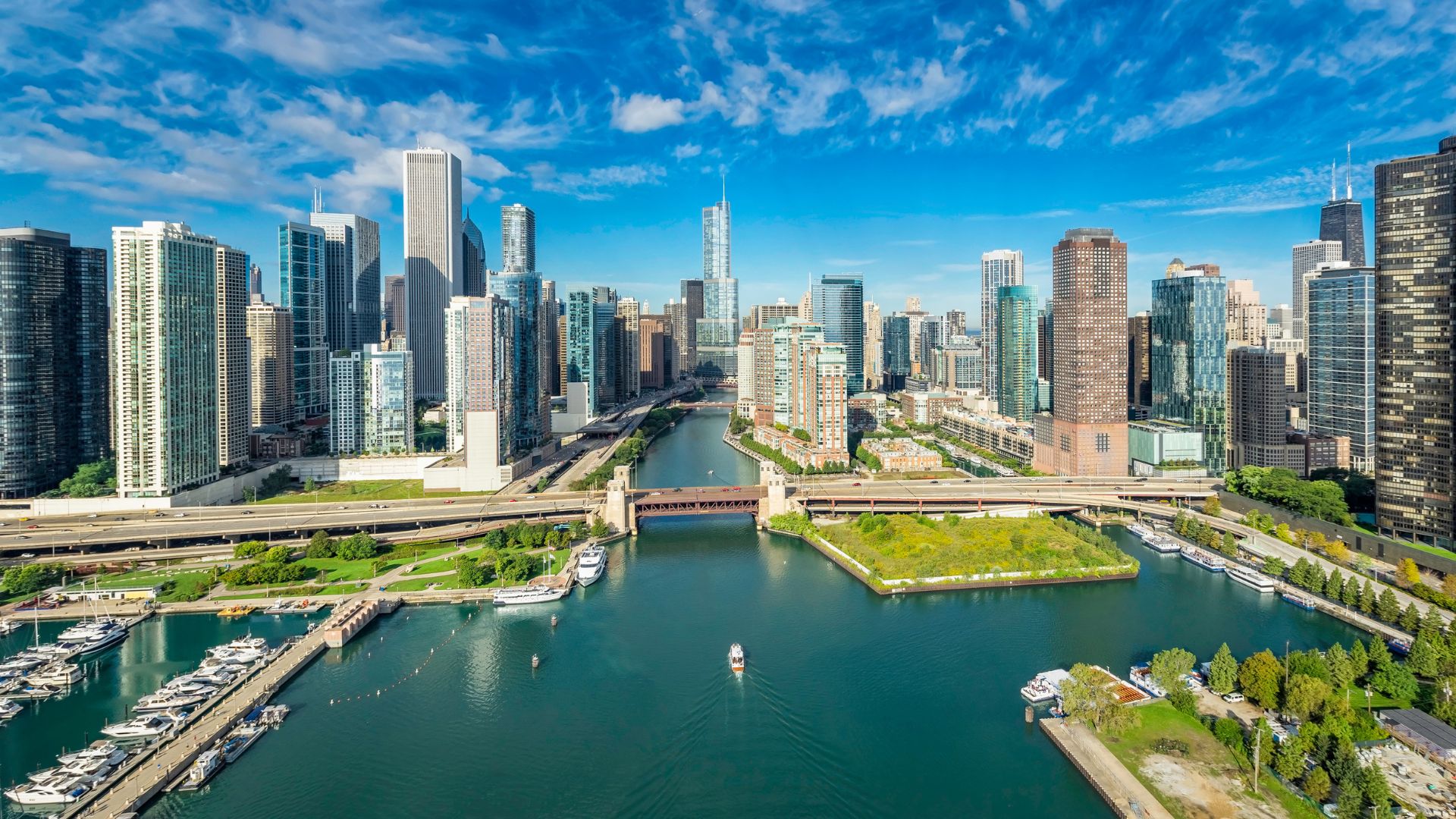 An aerial panorama of the modern Chicago skyline on a sunny day, showcasing a river with several bridges flowing between numerous high-rise skyscrapers and leading into a marina area in the foreground.