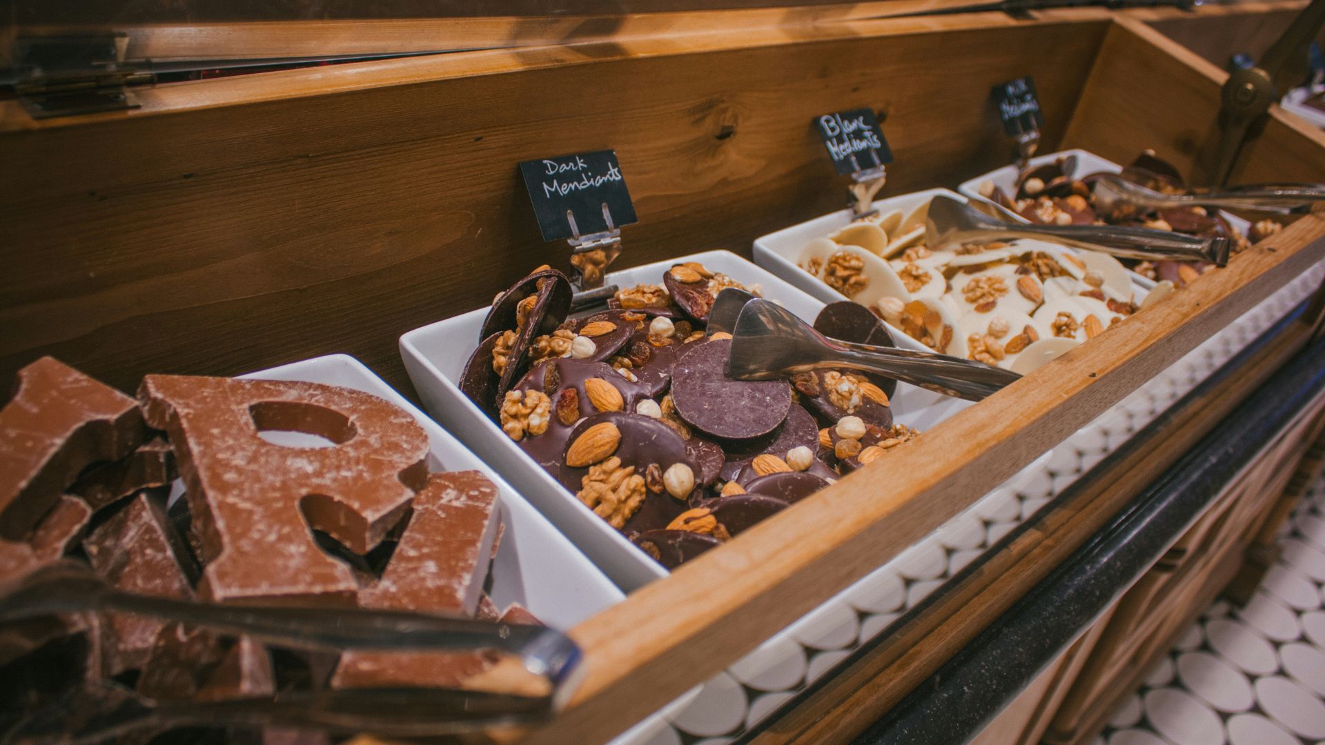 A close-up view of a self-serve counter display with assorted chocolates, dark mendiants, and white nut brittle in white containers.