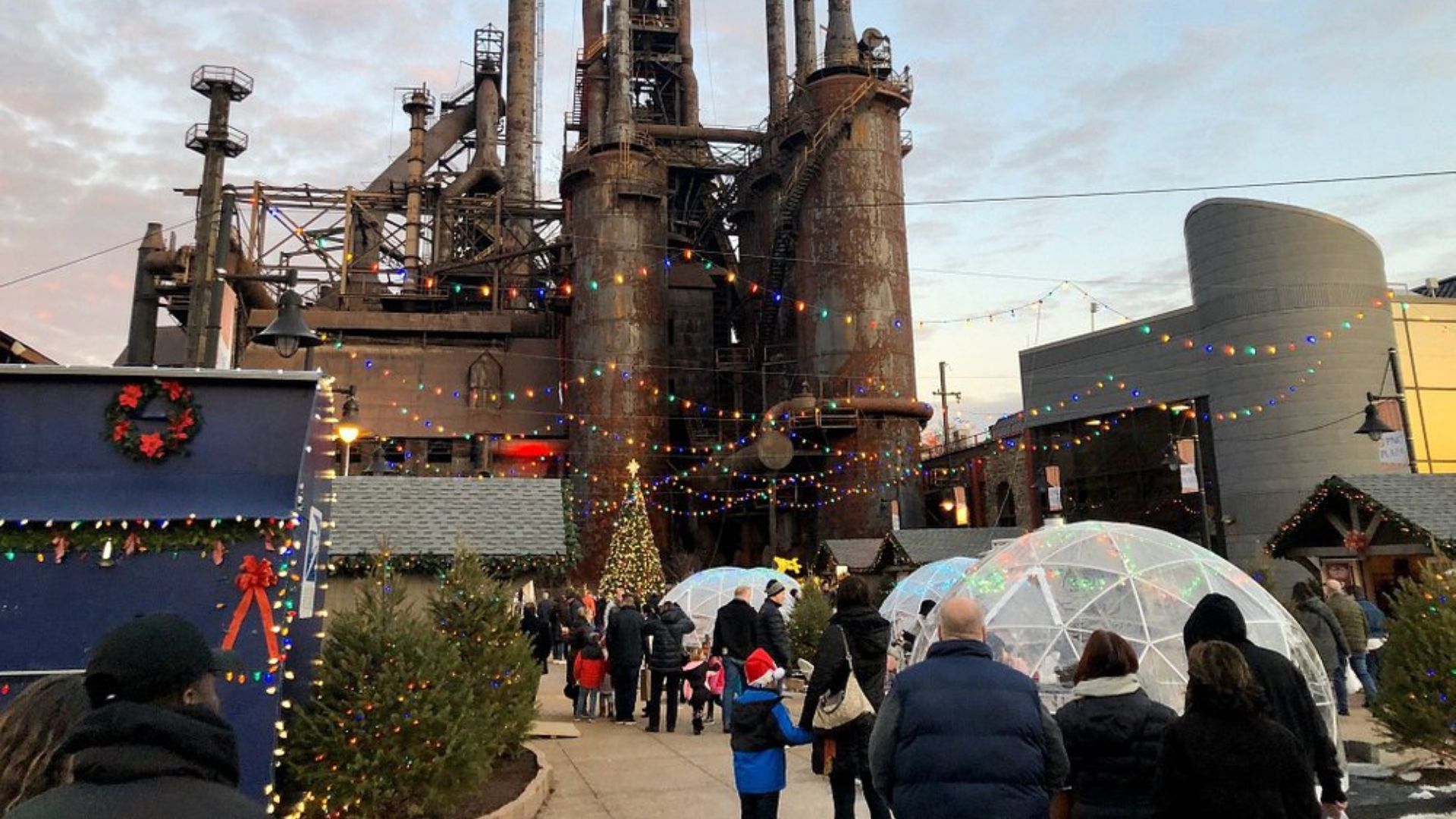 A busy outdoor Christmas market with people, stalls, and Christmas trees in the foreground, set against the backdrop of large, rusty industrial blast furnaces at dusk.