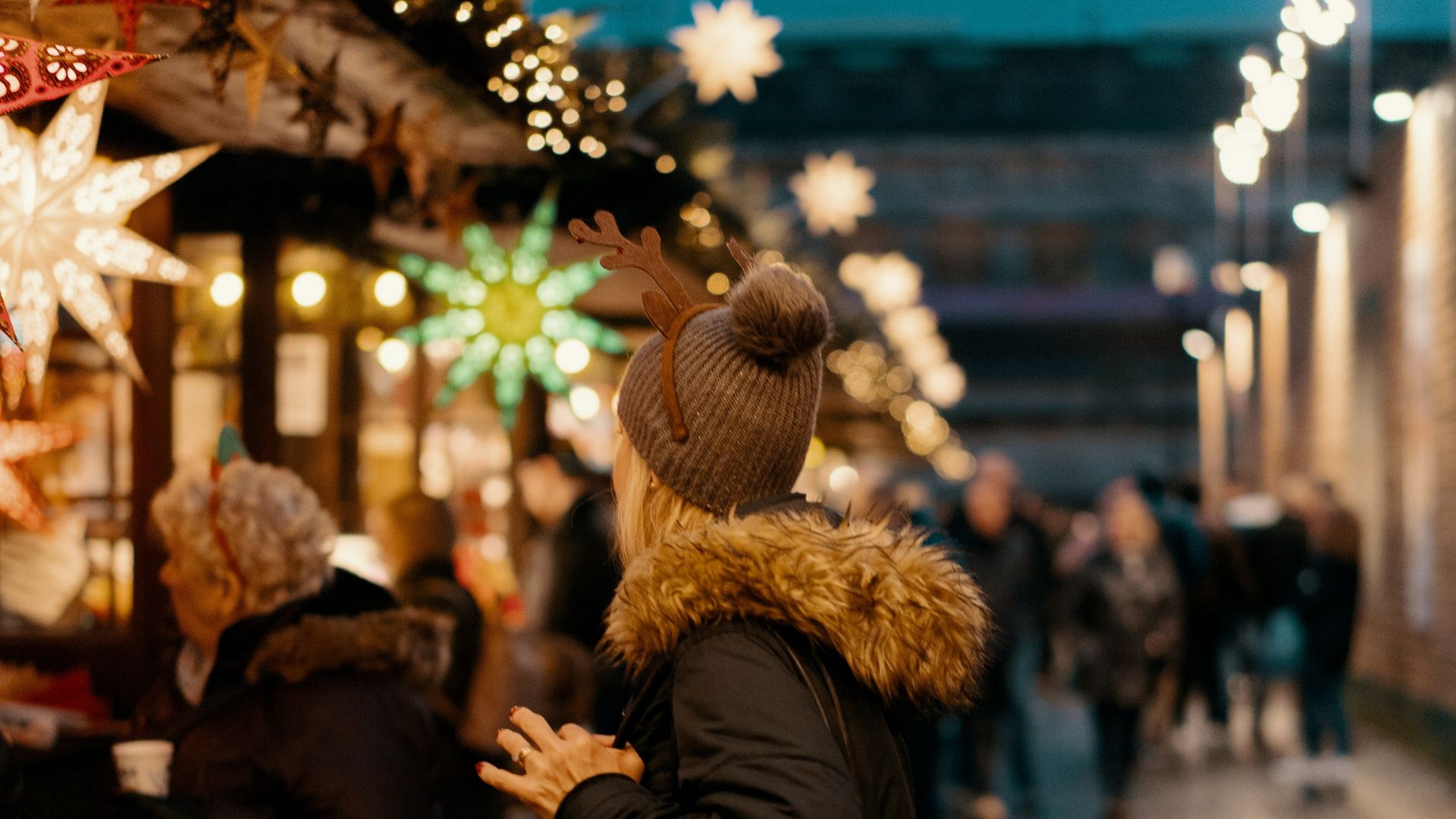 A person in a winter coat and hat with reindeer antlers holding a cup at a busy outdoor Christmas market at night, decorated with string lights and large star ornaments.