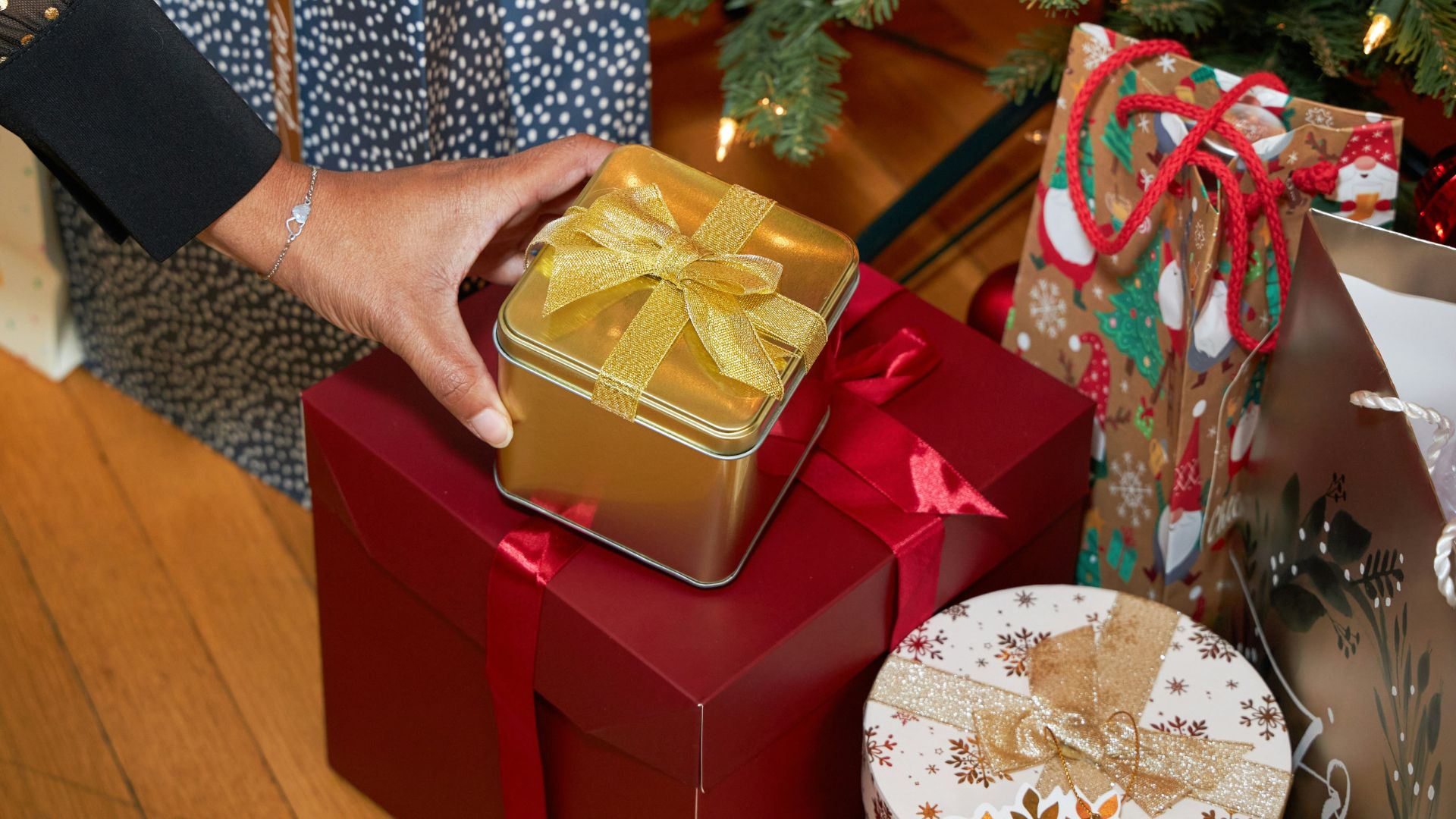 A hand selects a wrapped gift tin from a pile of Christmas presents in a Secret Santa scenario.